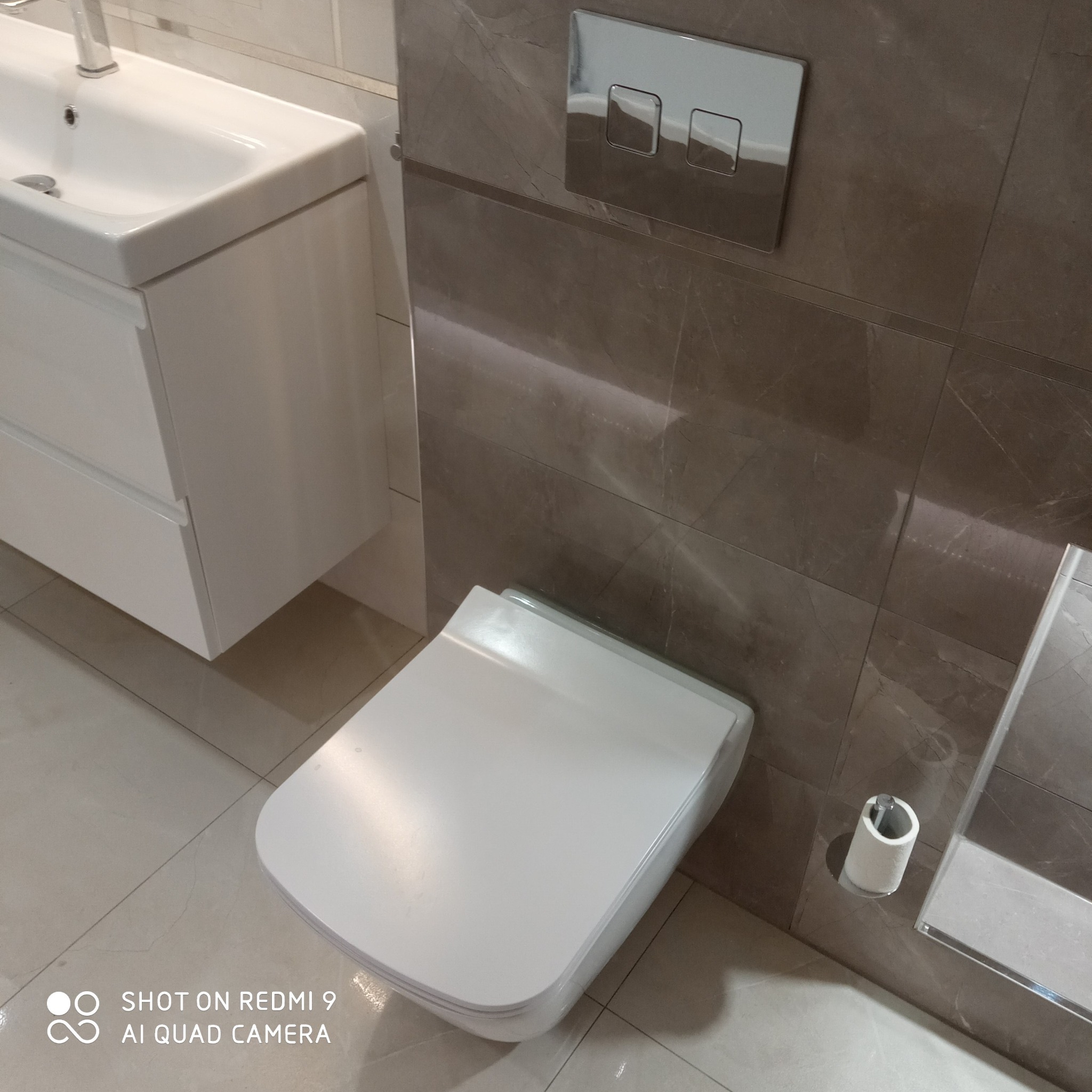 Nowoczesna, minimalistyczna łazienka z wiszącą toaletą, umywalką nablatową i brązowymi płytkami imitującymi marmur, widoczny chromowany przycisk spłuczki.