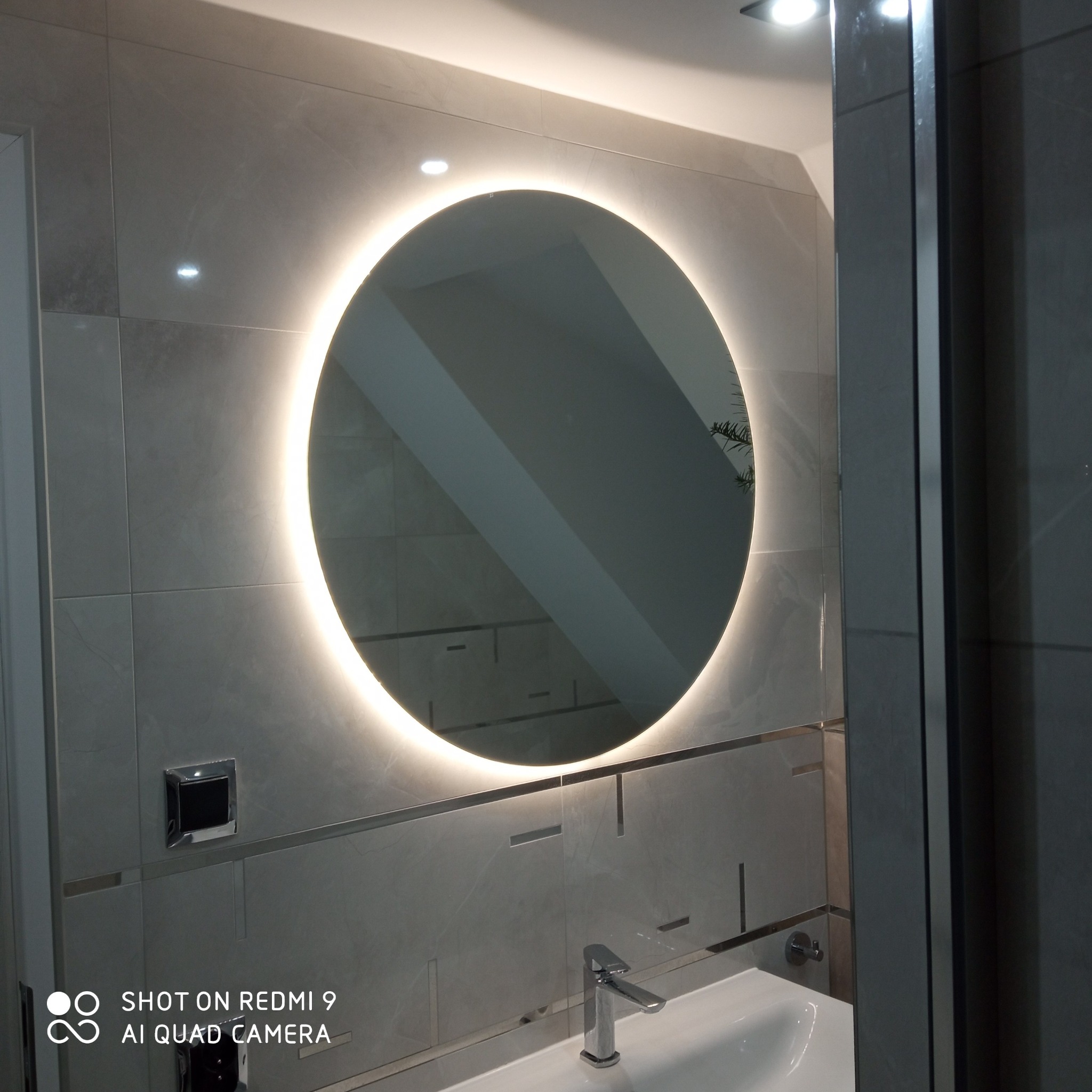 Nowoczesna łazienka z okrągłym lustrem podświetlanym LED, szare płytki imitujące marmur z dekoracyjnymi wstawkami, bateria umywalkowa w kolorze chrom, widoczny fragment kabiny prysznicowej.