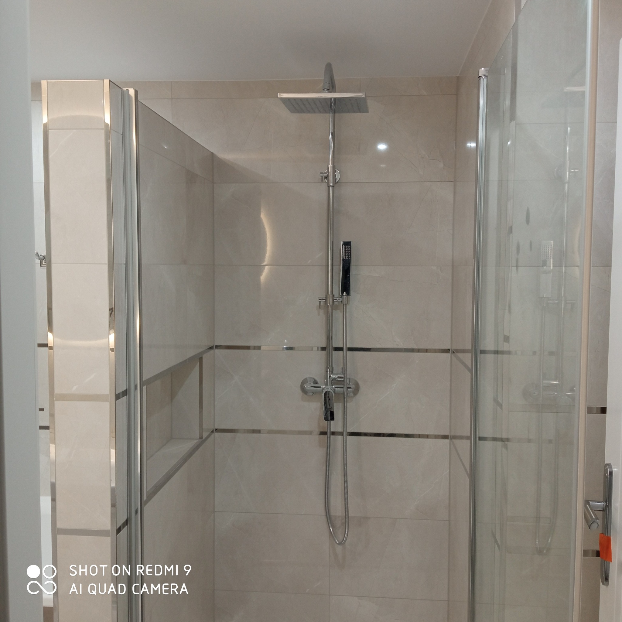 Kabina prysznicowa typu walk-in z szarymi płytkami imitującymi marmur, chromowanymi listwami dekoracyjnymi i deszczownicą, widok z przodu.