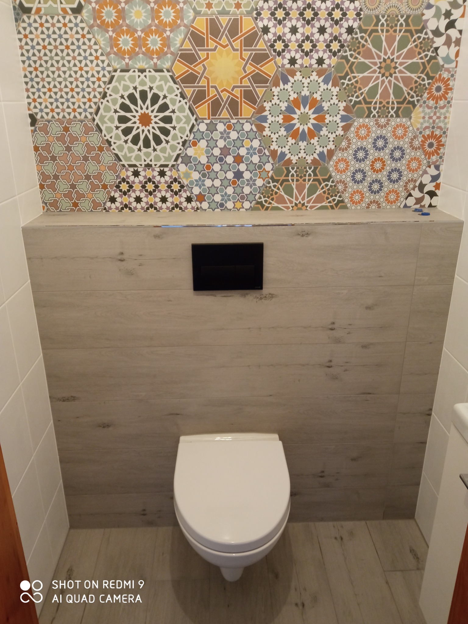 Nowoczesna toaleta podwieszana z czarnym przyciskiem spłukującym, obudowana płytami imitującymi drewno, nad którą umieszczono dekoracyjne heksagonalne płytki z motywami geometrycznymi.