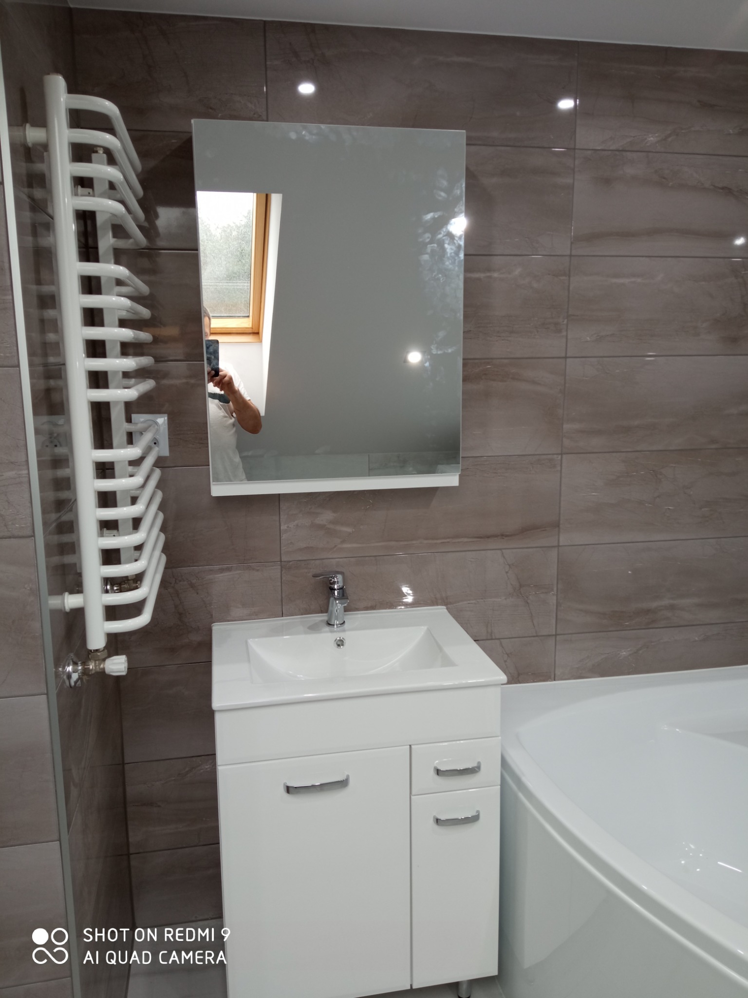 Nowoczesna łazienka na poddaszu z brązowymi płytkami imitującymi marmur, białą szafką pod umywalkę, grzejnikiem drabinkowym i narożną wanną; w lustrze odbija się okno dachowe i osoba robiąca zdjęcie.