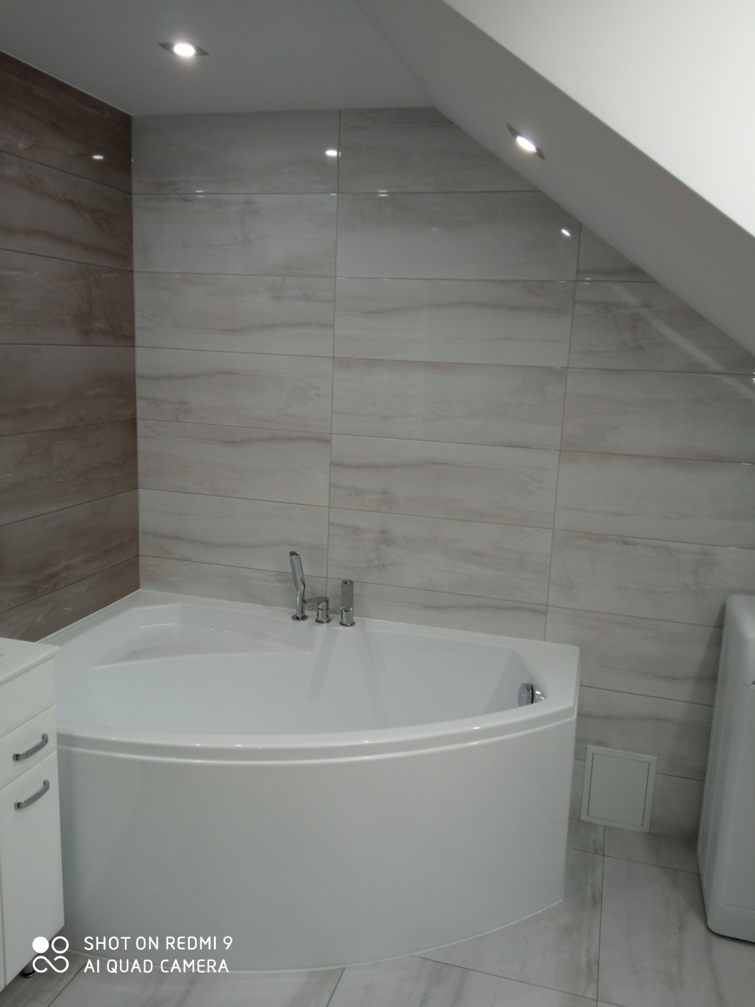 Nowoczesna łazienka na poddaszu z narożną wanną, wykończona płytkami imitującymi marmur, oświetlona punktowymi lampami sufitowymi.