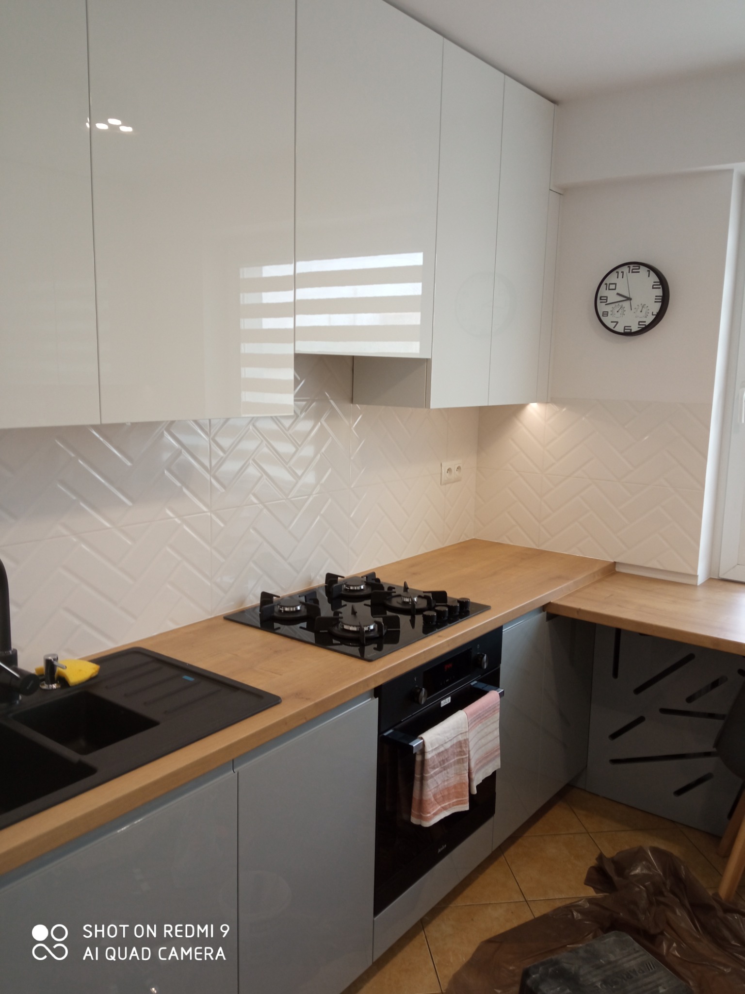 Nowoczesna kuchnia z białymi szafkami, drewnianym blatem i białymi płytkami ułożonymi w jodełkę na ścianie, czarny zlew i płyta grzewcza, zegar na ścianie.