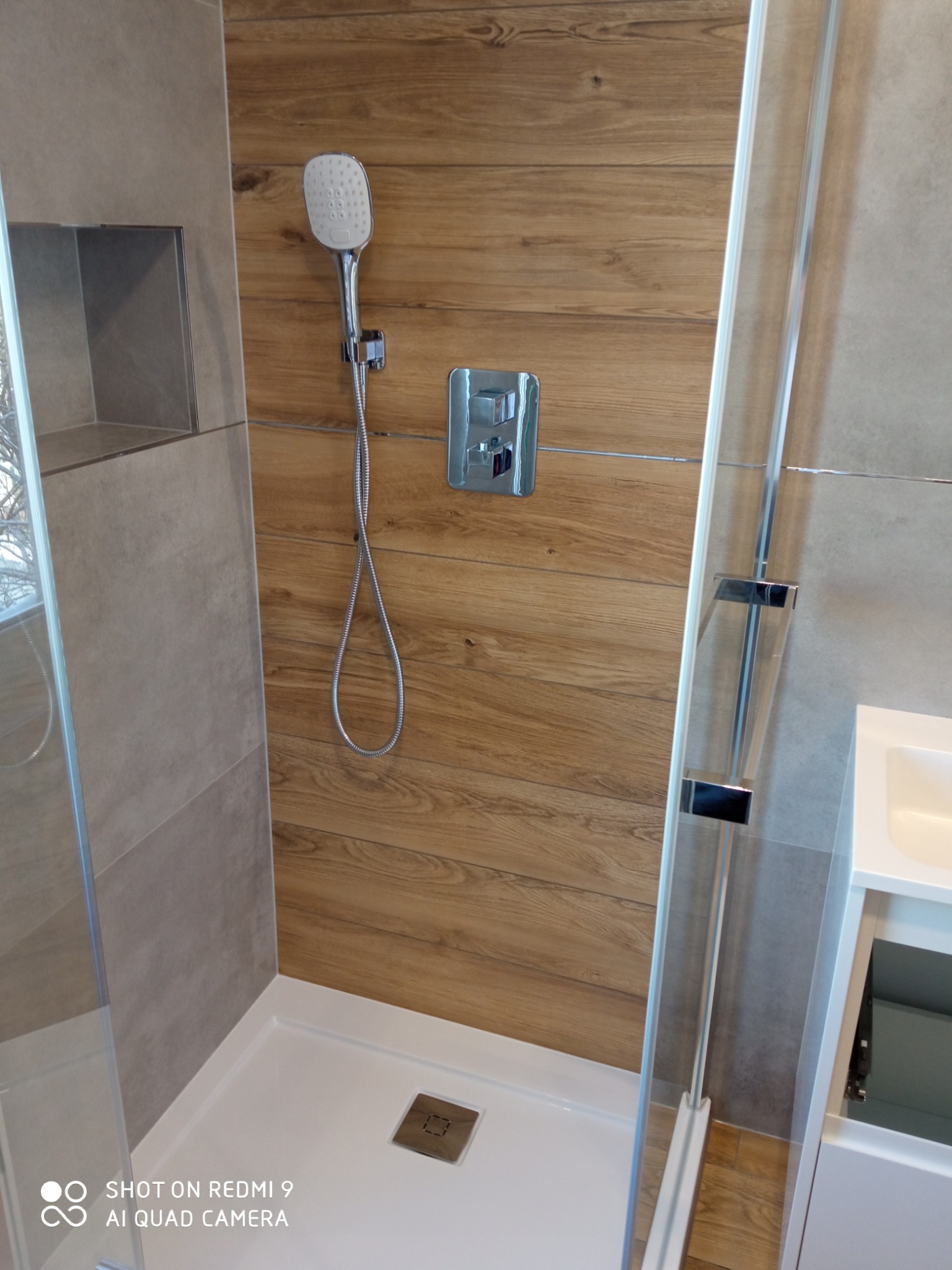 Nowoczesna kabina prysznicowa z deszczownicą i drewnopodobnymi płytkami w Szczytnie, widoczny odpływ liniowy w brodziku i szklane drzwi przesuwne.