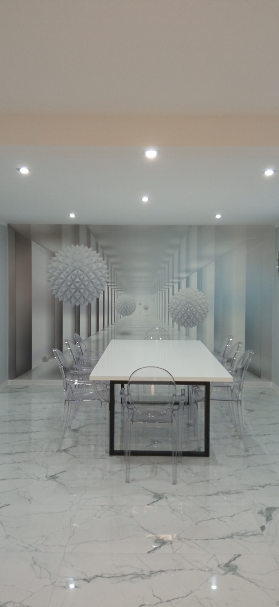 Nowoczesna jadalnia z białym stołem i przezroczystymi krzesłami na podłodze z płytek imitujących marmur, ściana z fototapetą z motywem geometrycznym.