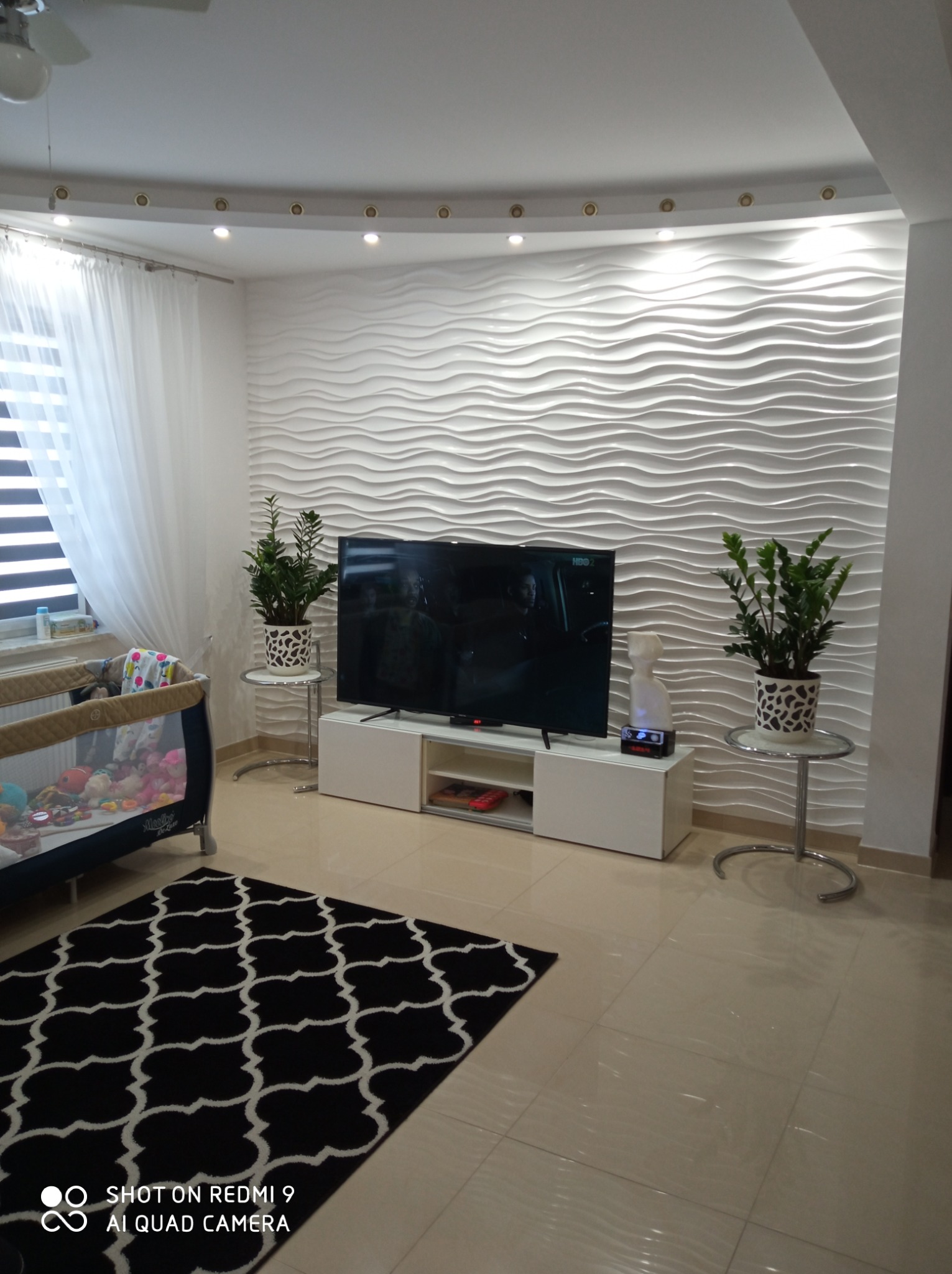 Nowoczesny salon z dekoracyjną ścianą 3D w faliste wzory, podświetlanym sufitem, beżową podłogą z płytek, czarnym dywanem w białe wzory, białymi meblami RTV, telewizorem, roślinami doniczkowymi...