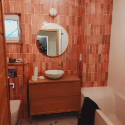 Wykończona łazienka z terakotową glazurą ułożoną pionowo, okrągłe lustro z podświetleniem, drewniana szafka pod umywalką i widoczna wanna, w lustrze odbicie osoby robiącej zdjęcie.