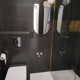 Nowoczesna łazienka z czarnymi płytkami imitującymi skórę, owalnym lustrem z podświetleniem i białą armaturą, widoczna kabina prysznicowa ze złotymi akcentami.