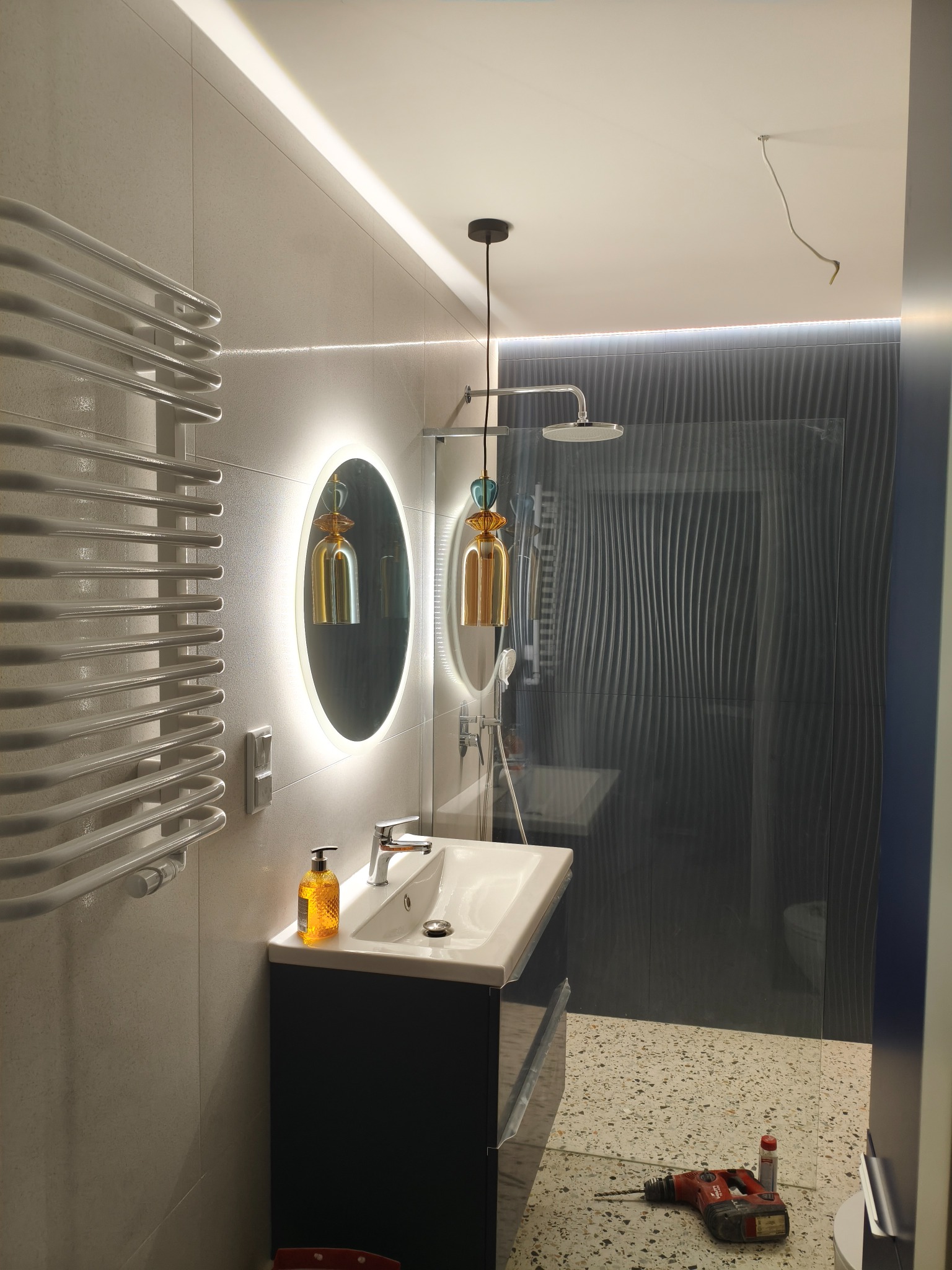 Nowoczesna łazienka z owalnym lustrem LED, grzejnikiem drabinkowym i kabiną prysznicową z falistą ścianą. Na podłodze wiertarka i akcesoria.