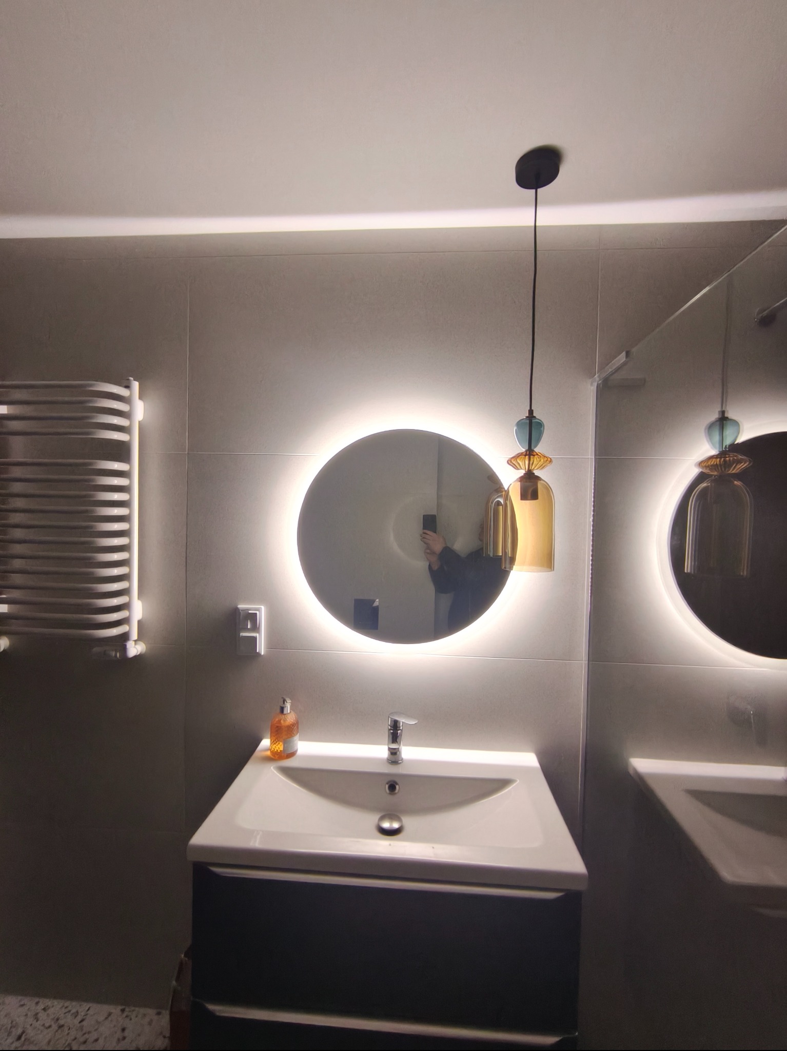 Nowoczesna łazienka z okrągłym lustrem podświetlanym LED, wiszącą lampą dekoracyjną i grzejnikiem drabinkowym. Minimalistyczny design, szare płytki.