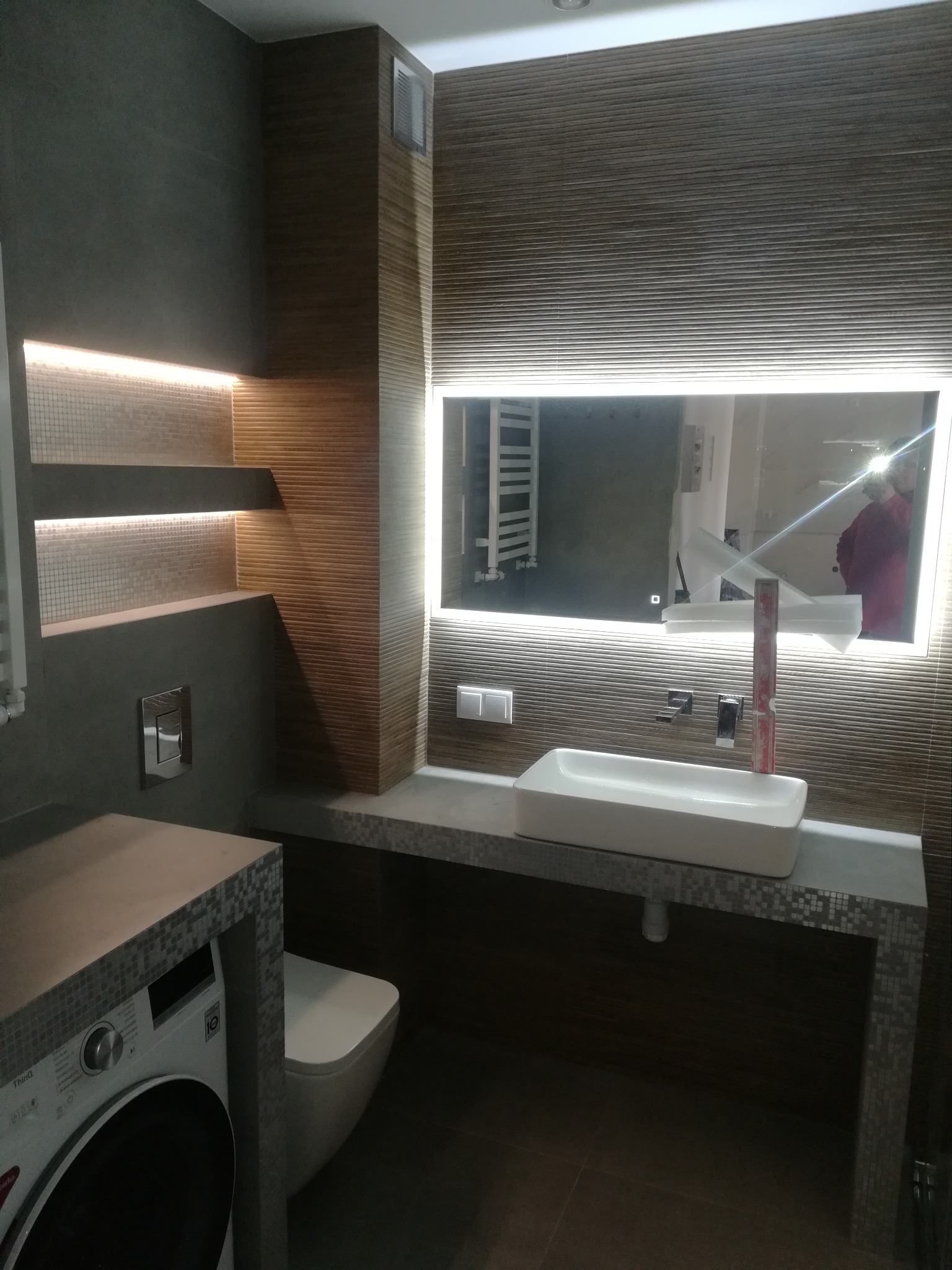 Nowoczesna łazienka z podświetlanymi półkami, lustrem z oświetleniem LED, umywalką nablatową i pralką obudowaną mozaiką.