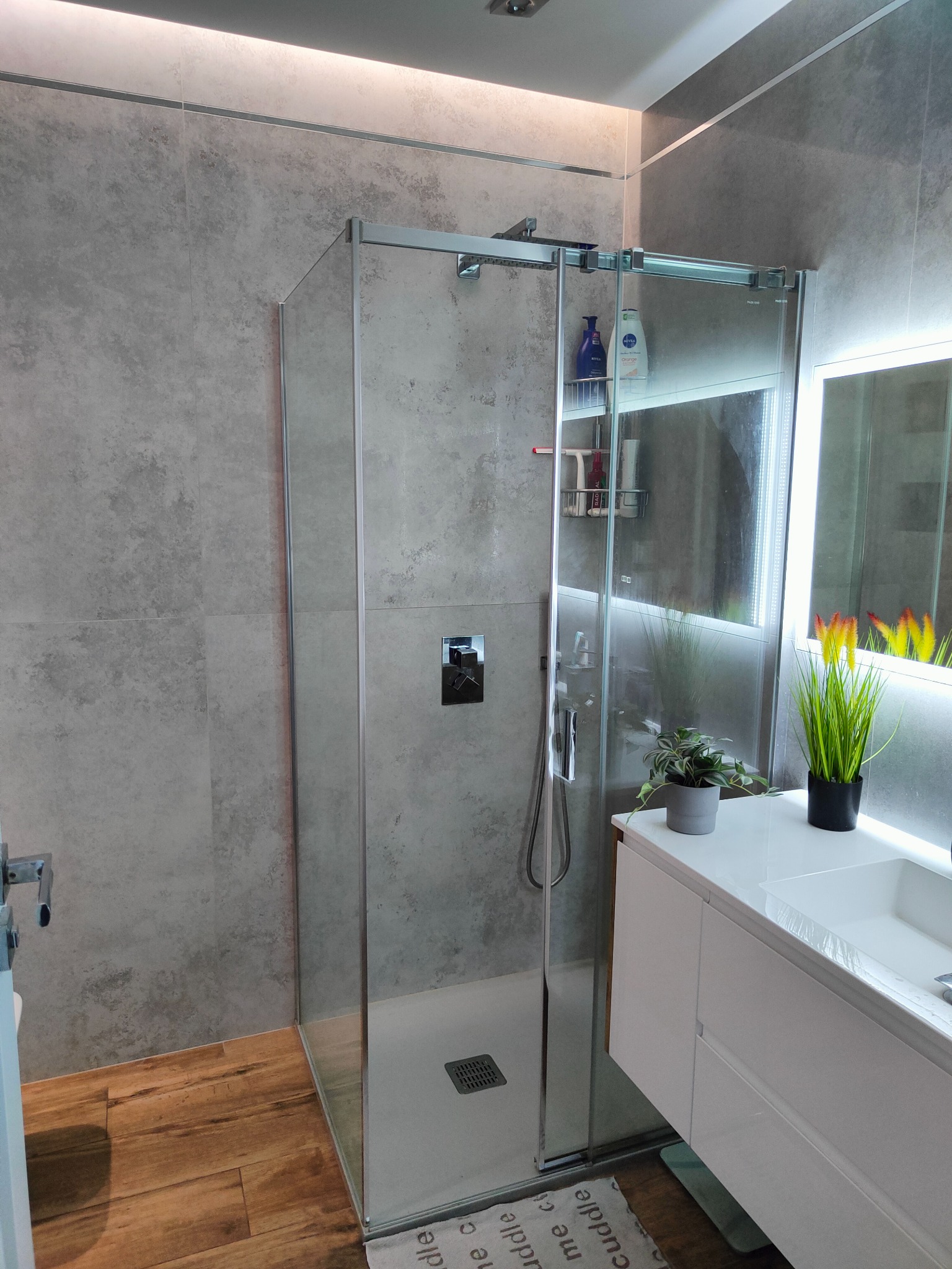 Nowoczesna kabina prysznicowa z szarymi płytkami imitującymi beton i drewnianą podłogą w łazience we Wrocławiu, z podświetlanym lustrem i roślinami na blacie.