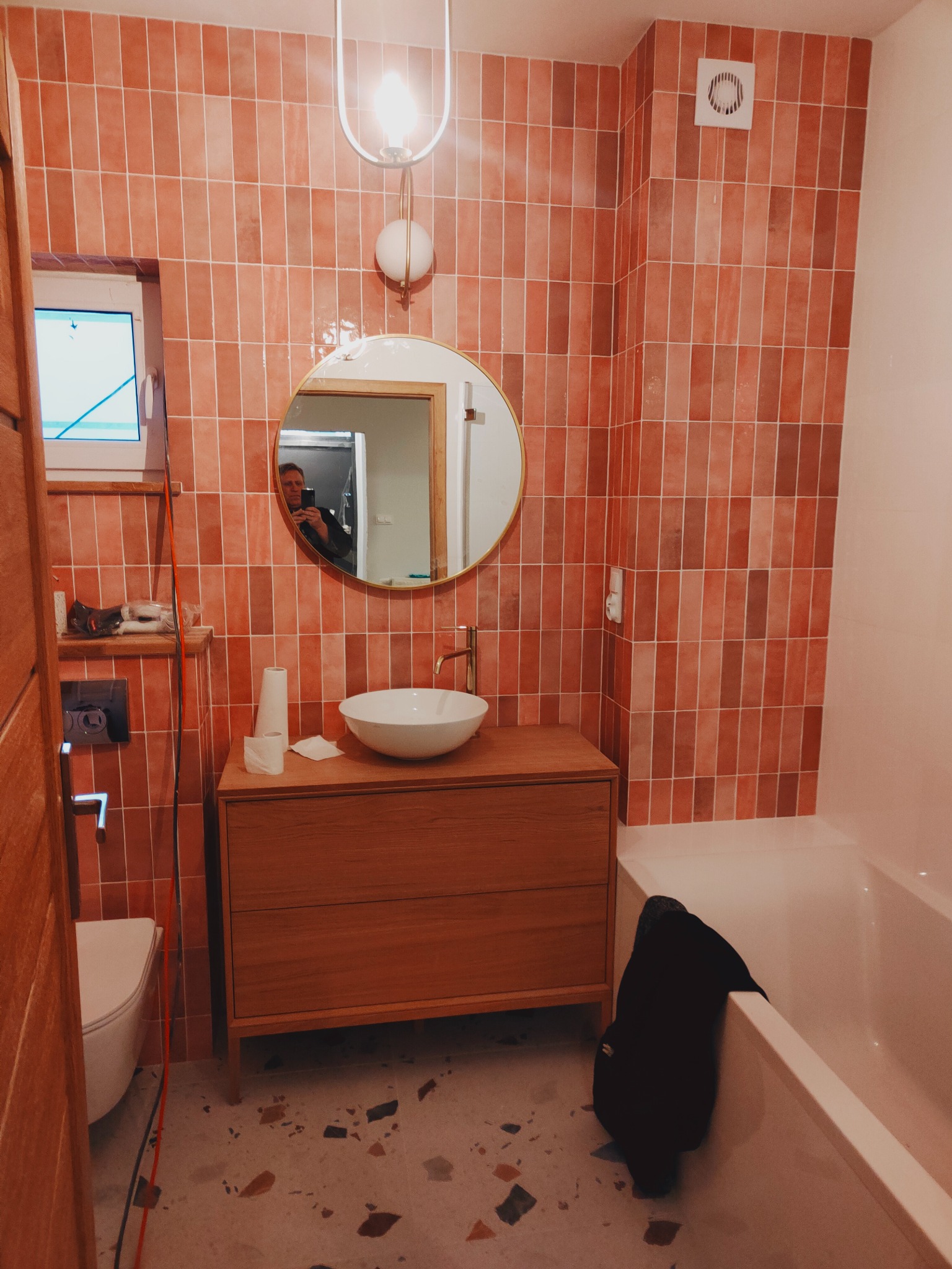 Wykończona łazienka z terakotową glazurą ułożoną pionowo, okrągłe lustro z podświetleniem, drewniana szafka pod umywalką i widoczna wanna, w lustrze odbicie osoby robiącej zdjęcie.