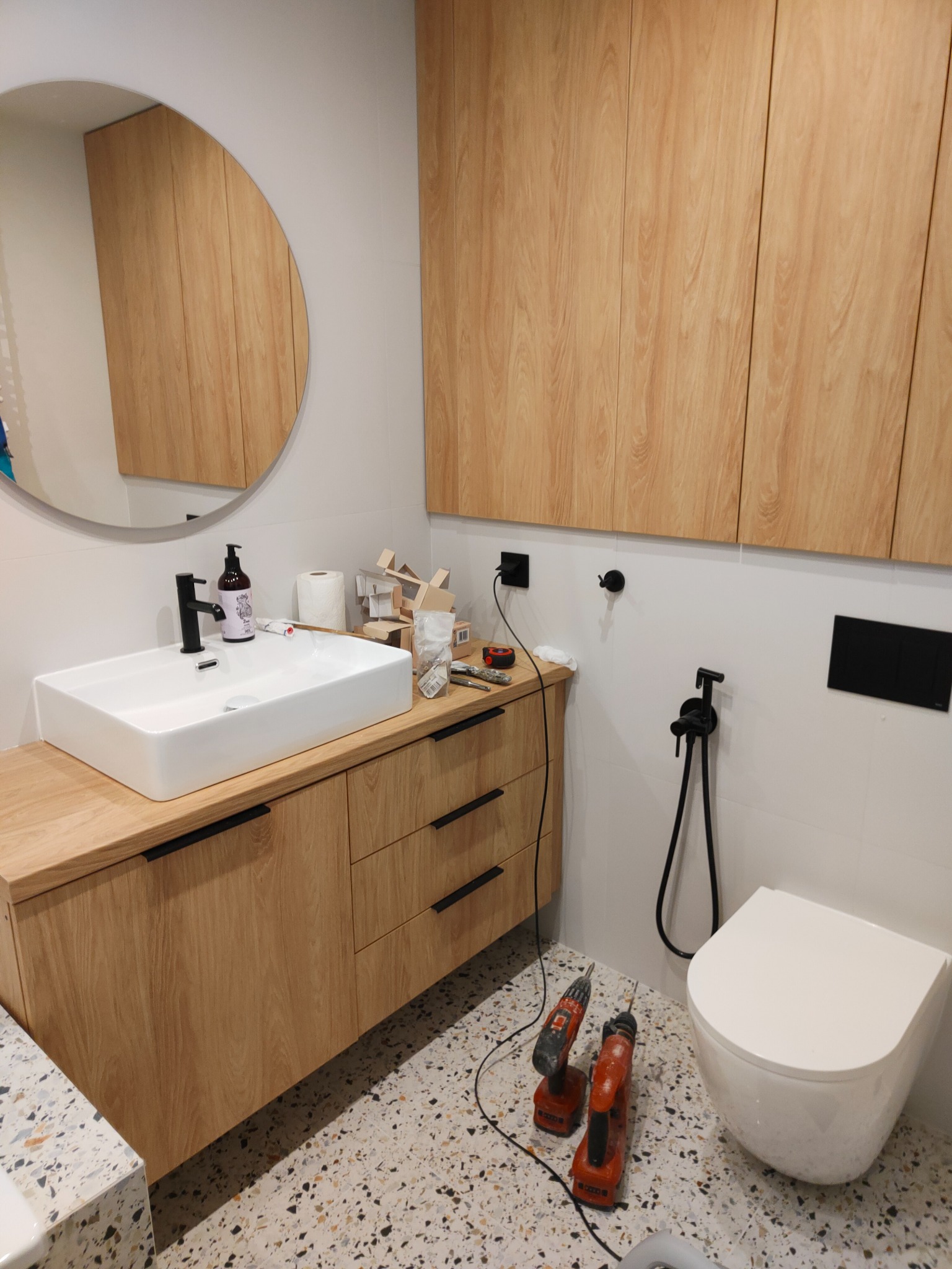 Montaż umywalki nablatowej, wiszącej toalety i armatury w łazience z mozaikową podłogą, widoczne wiertarki na podłodze i akcesoria montażowe na blacie.