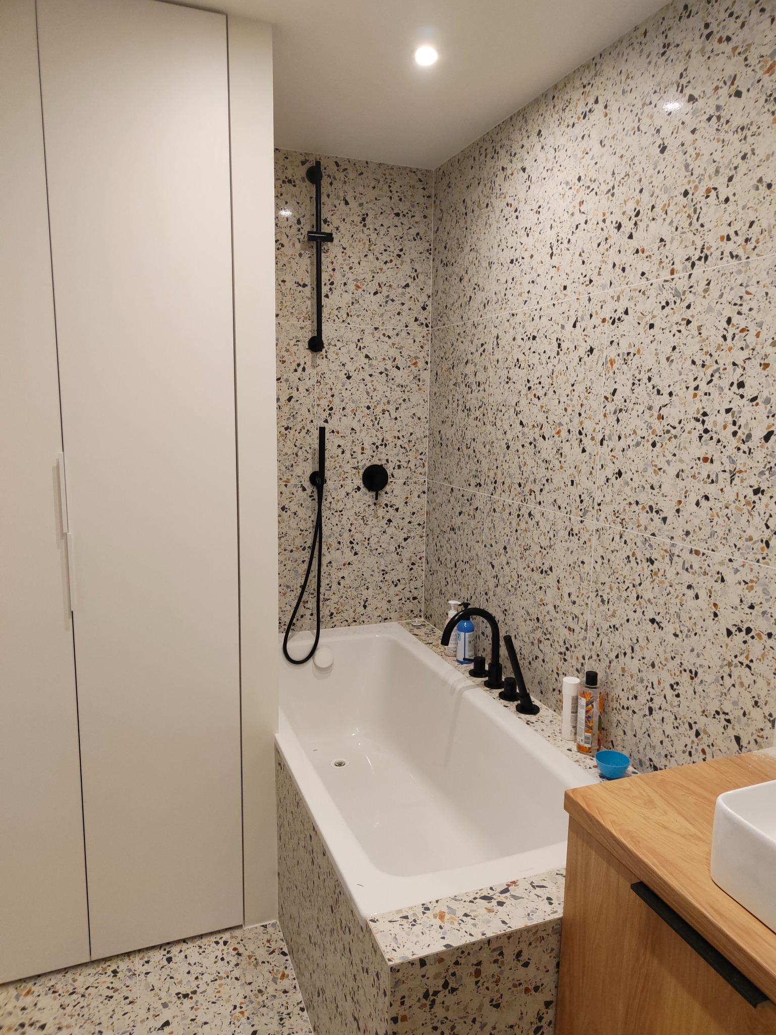 Biała wanna z czarną armaturą i prysznicem na tle ściany i podłogi wykończonej płytkami lastryko w łazience, obok szafka z umywalką i wysoka szafka.