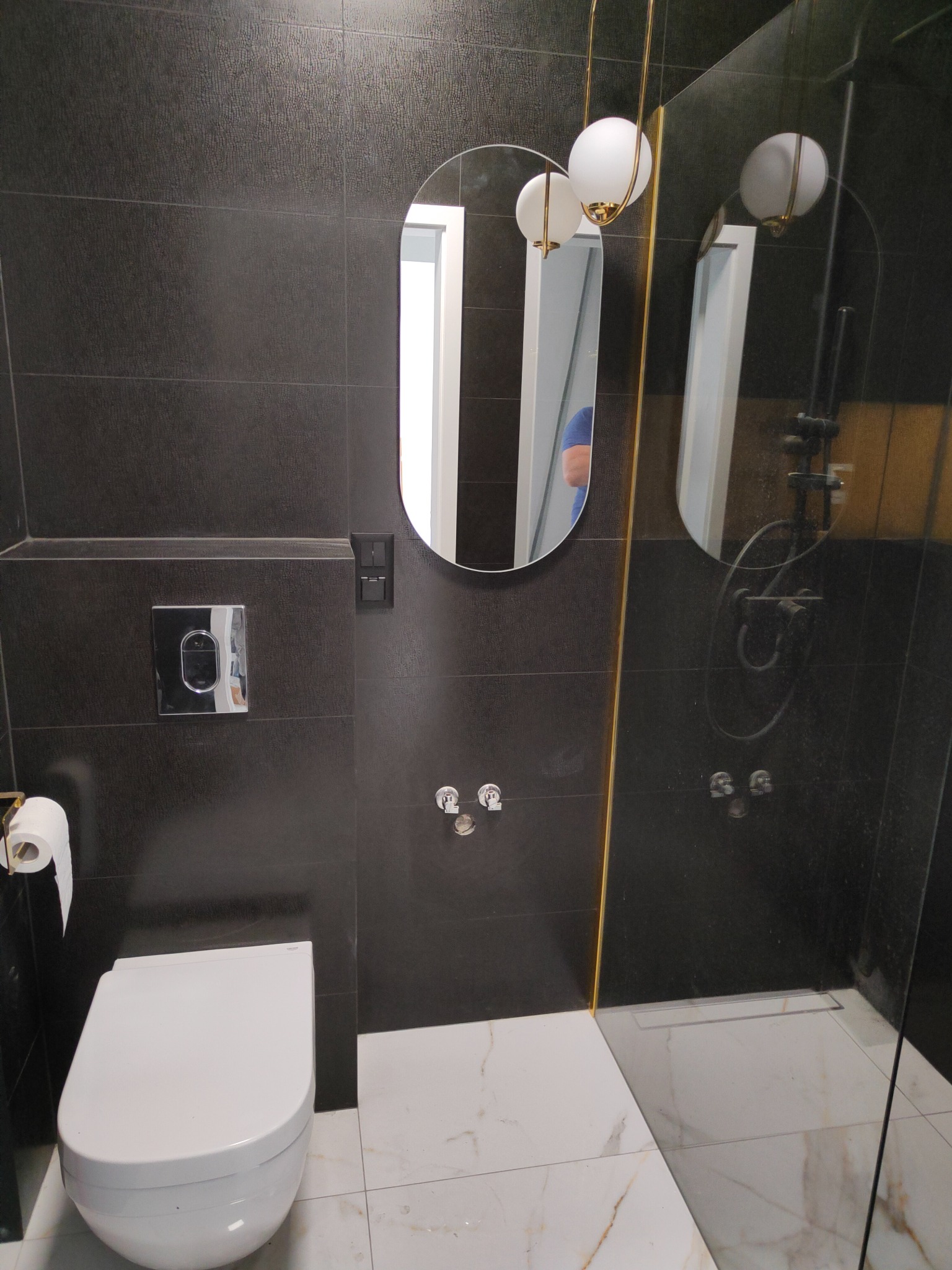 Nowoczesna łazienka z czarnymi płytkami imitującymi skórę, owalnym lustrem z podświetleniem i białą armaturą, widoczna kabina prysznicowa ze złotymi akcentami.