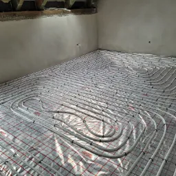 Białe rury ogrzewania podłogowego ułożone spiralnie na srebrnej folii z czerwoną kratką, widok z góry w surowym stanie, poddasze z oknem dachowym.