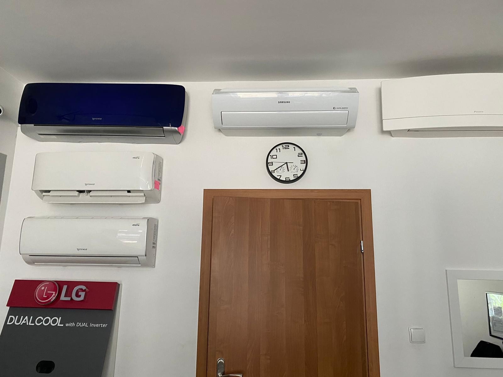 Wystawa klimatyzatorów różnych marek (Rotenso, Samsung, Daikin, LG) zamontowanych na białej ścianie nad drewnianymi drzwiami i obok lustra, z widocznym zegarem ściennym.