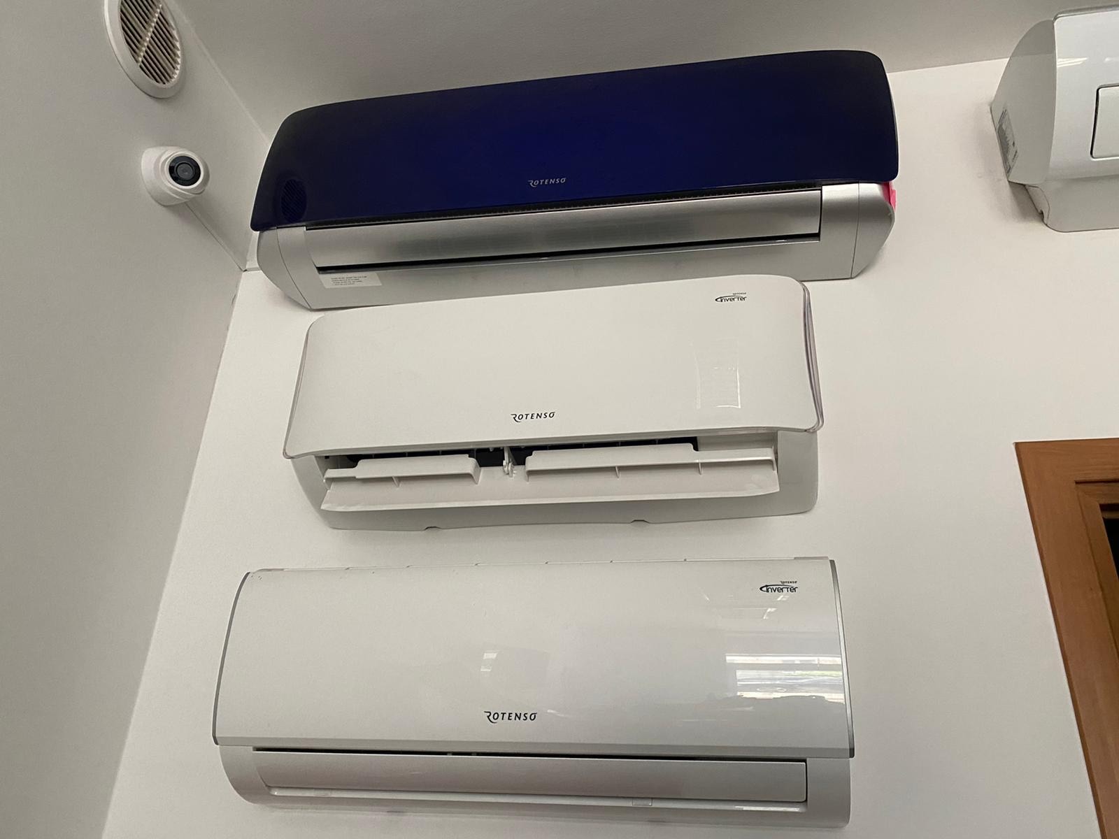 Trzy jednostki klimatyzacyjne Rotenso zamontowane na białej ścianie, górna w kolorze granatowym, środkowa z otwartymi klapami, widoczna kamera monitoringu i kratka wentylacyjna