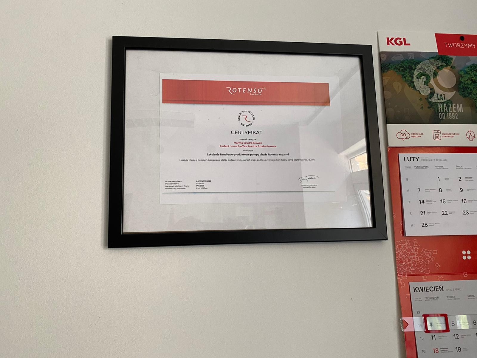 Oprawiony certyfikat Rotenso, potwierdzający ukończenie szkolenia handlowo-produktowego pomp ciepła Aquami, wiszący na ścianie obok kalendarza firmowego KGL.