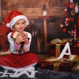 Uśmiechnięta dziewczynka w czapce Mikołaja siedzi na dywanie obok prezentów z literami tworzącymi słowo SANTA, na tle bożonarodzeniowej scenerii z drewnianym konikiem na biegunach.
