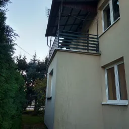 Zewnętrzny widok balkonu z poziomą, metalową konstrukcją zabezpieczającą, zadaszeniem i anteną na dachu, przylegającego do jasnej ściany budynku z białymi oknami, otoczonego zielenią.