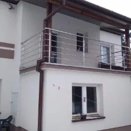 Balkon z nowoczesną, poziomą balustradą ze stali nierdzewnej, widoczny z zewnątrz budynku z elementami wykończenia w kolorze brązowym.