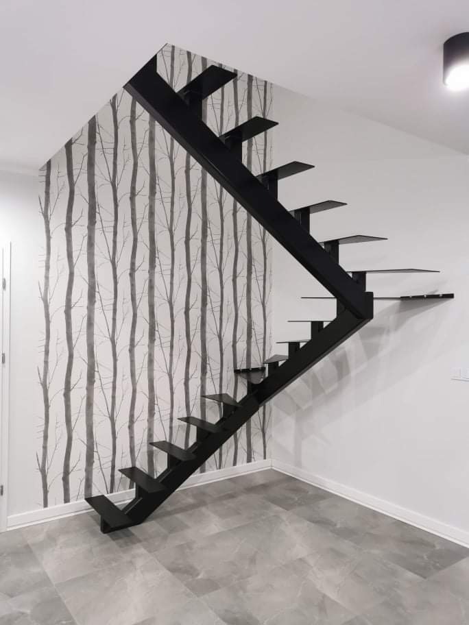 Nowoczesne, czarne schody o minimalistycznej konstrukcji, zamontowane przy ścianie z tapetą w motywie drzew, na tle szarej podłogi.