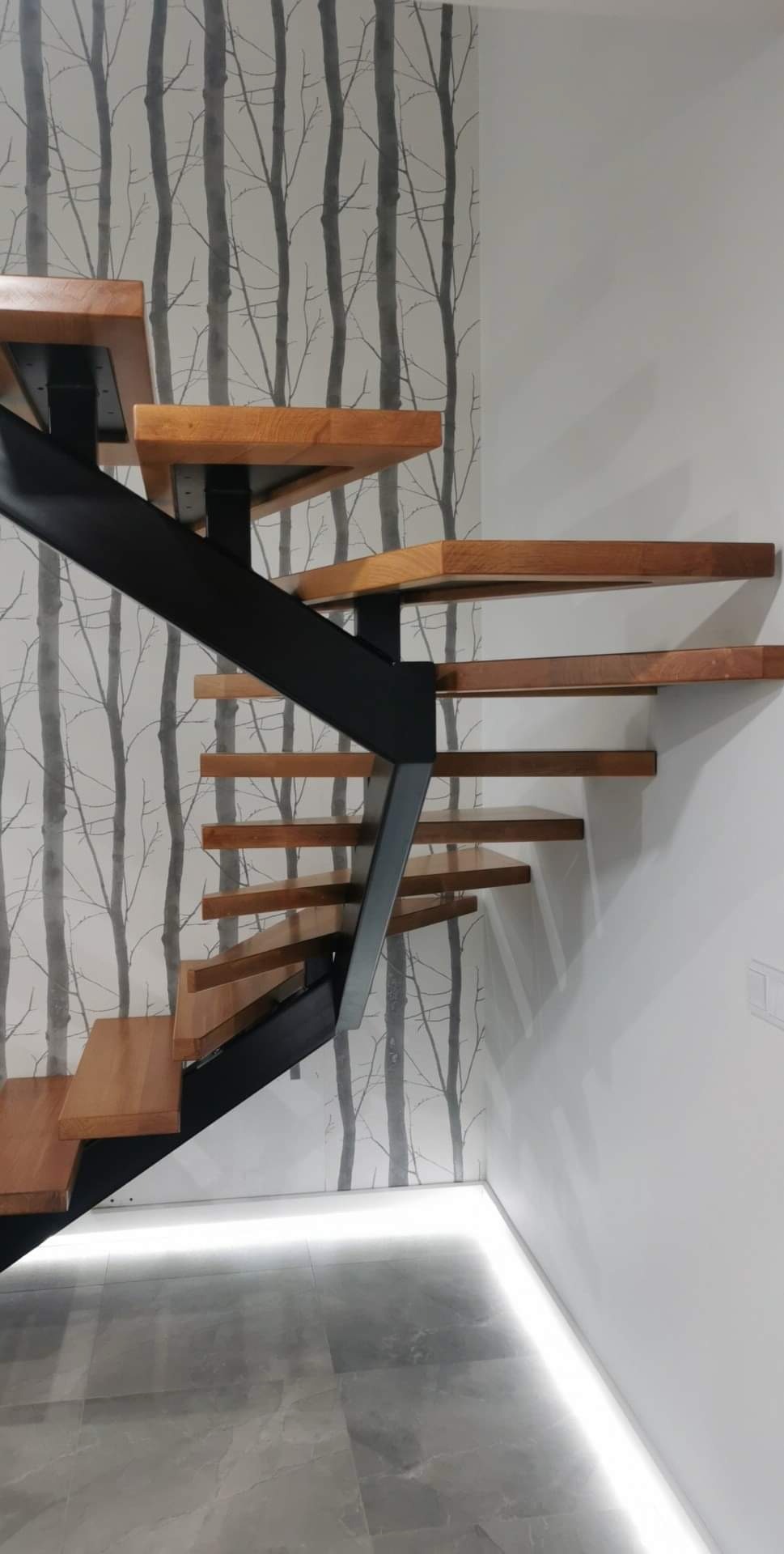 Nowoczesne schody z drewnianymi stopniami i czarną metalową konstrukcją, podświetlenie LED u dołu, na tle ściany z motywem drzew.
