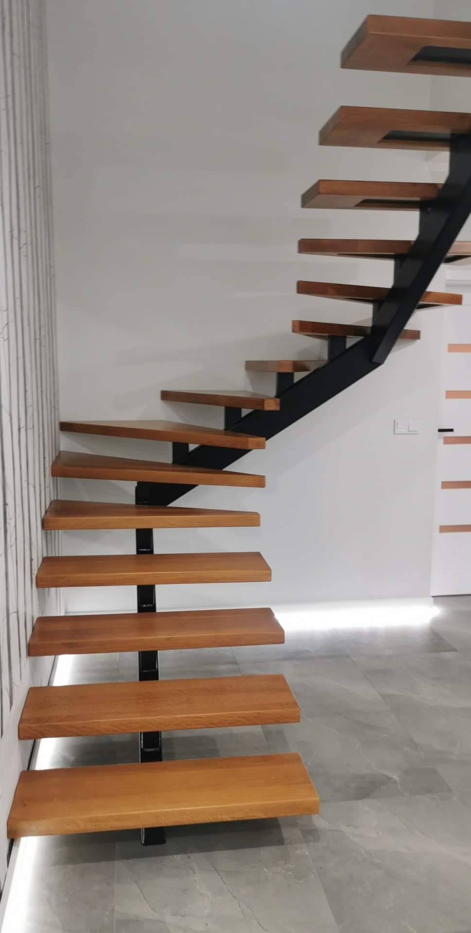 Nowoczesne schody z drewnianymi stopniami i czarną, metalową konstrukcją nośną, zamontowane przy ścianie z dekoracyjną tapetą w pionowe pasy.