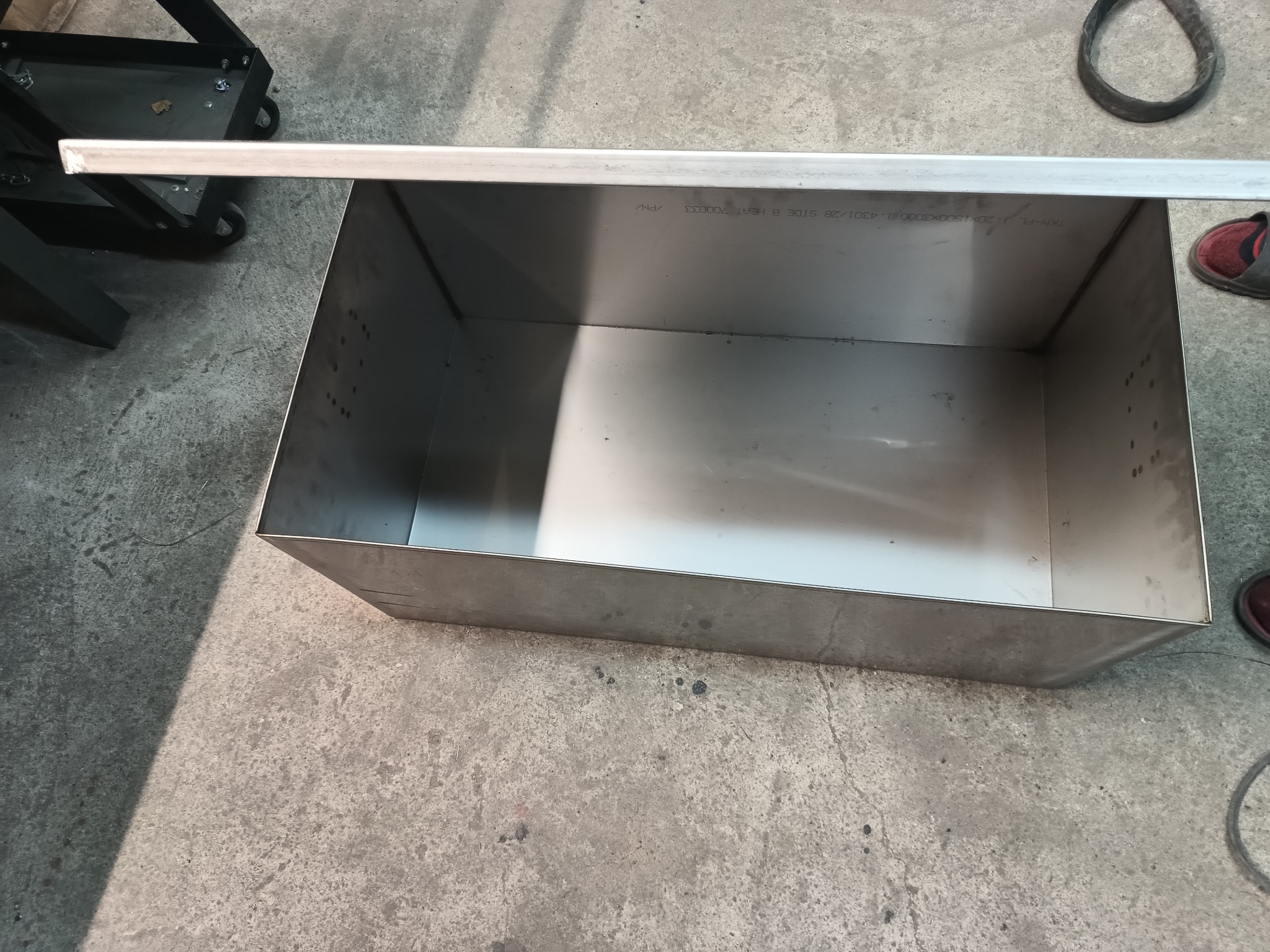 Metalowa skrzynia z nierdzewnej stali w warsztacie, widoczne oznaczenia techniczne na wewnętrznej ściance, obok fragment podłogi, w tle wózek warsztatowy i fragment obuwia.