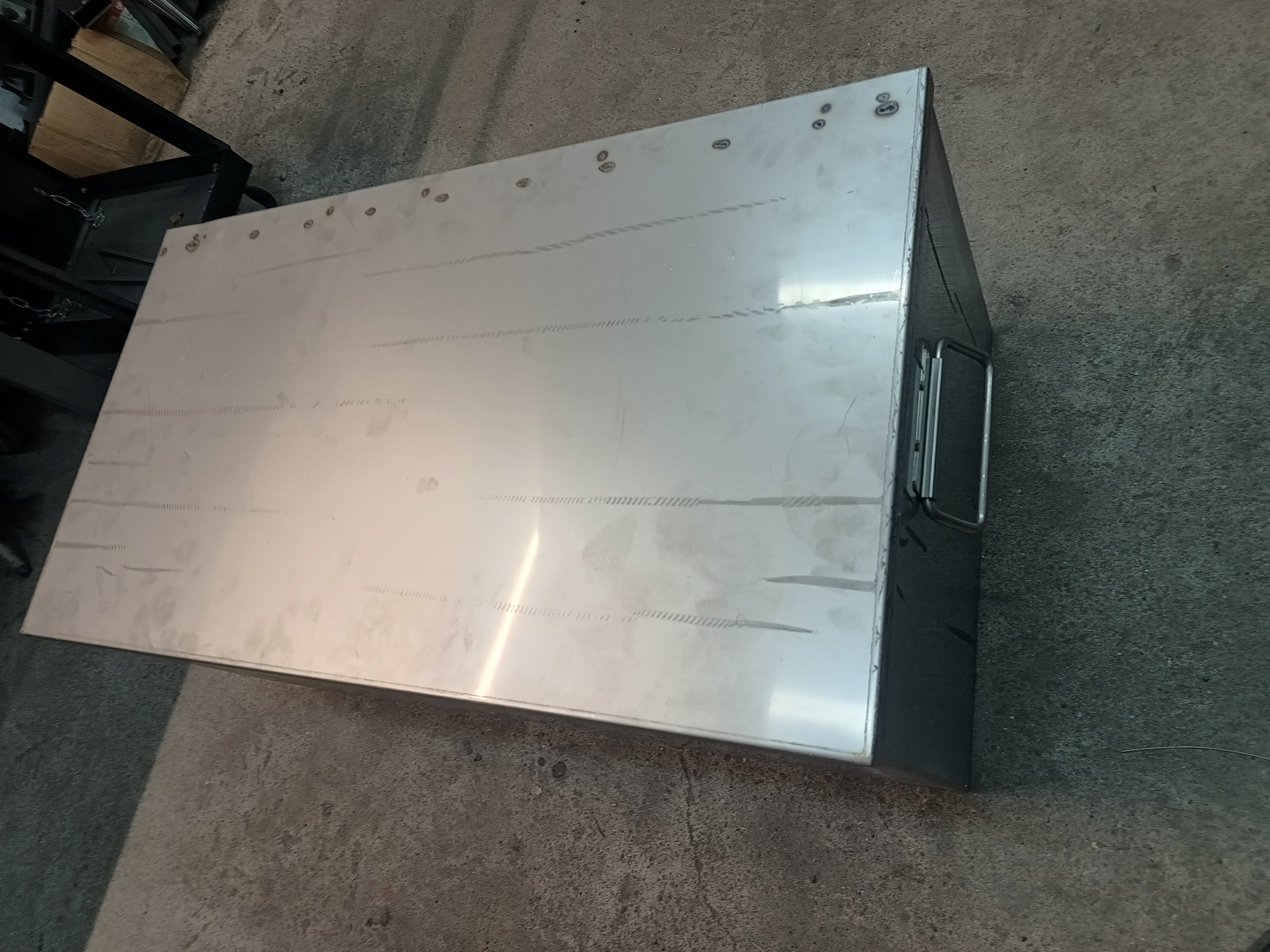 Metalowa skrzynia z uchwytami, wykonana z blachy, z widocznymi nitami i śladami po szlifowaniu, leżąca na betonowej podłodze.