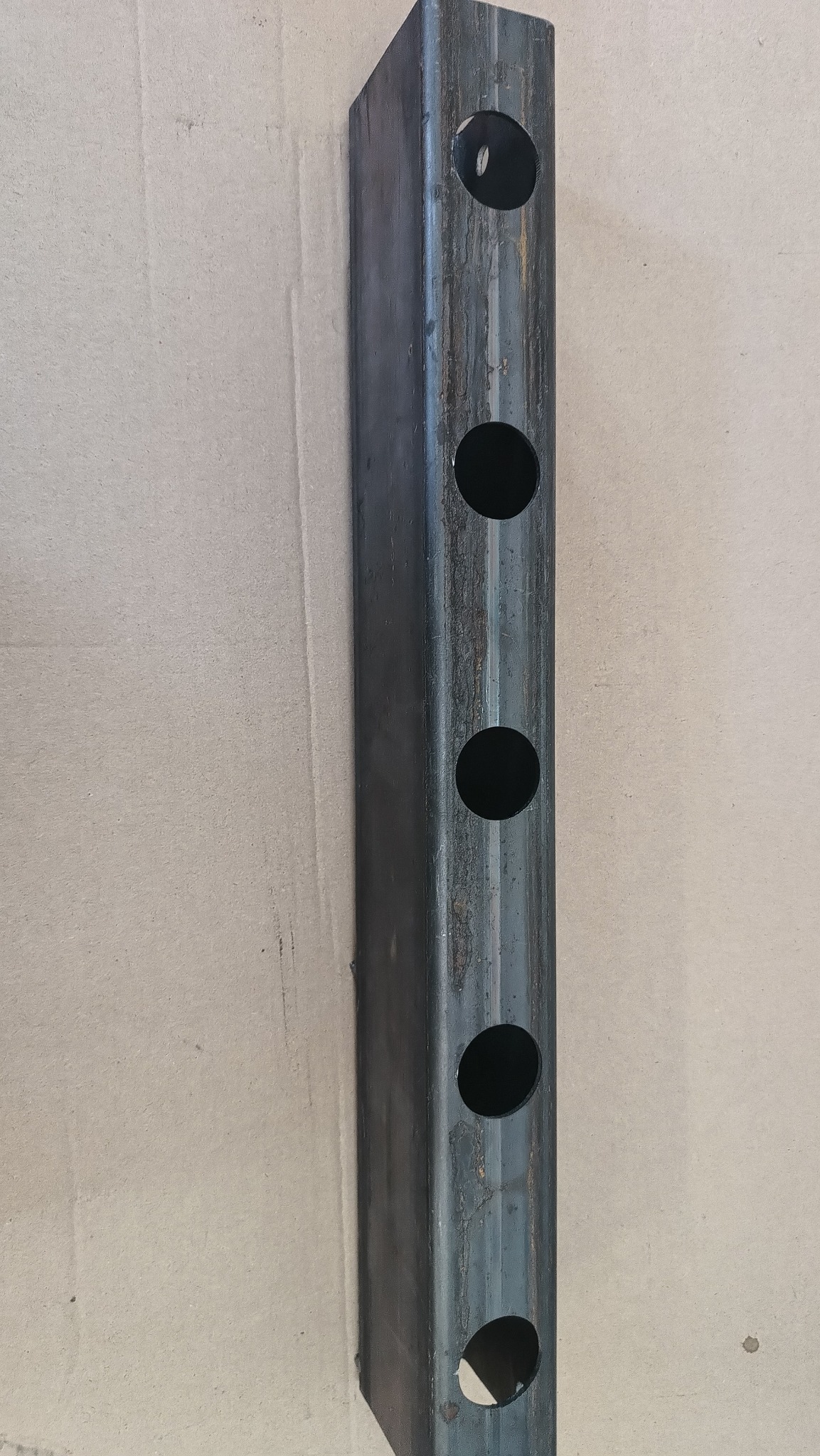 Czworokątny metalowy profil z otworami, surowy stan, widoczne ślady obróbki i rdzawy nalot.