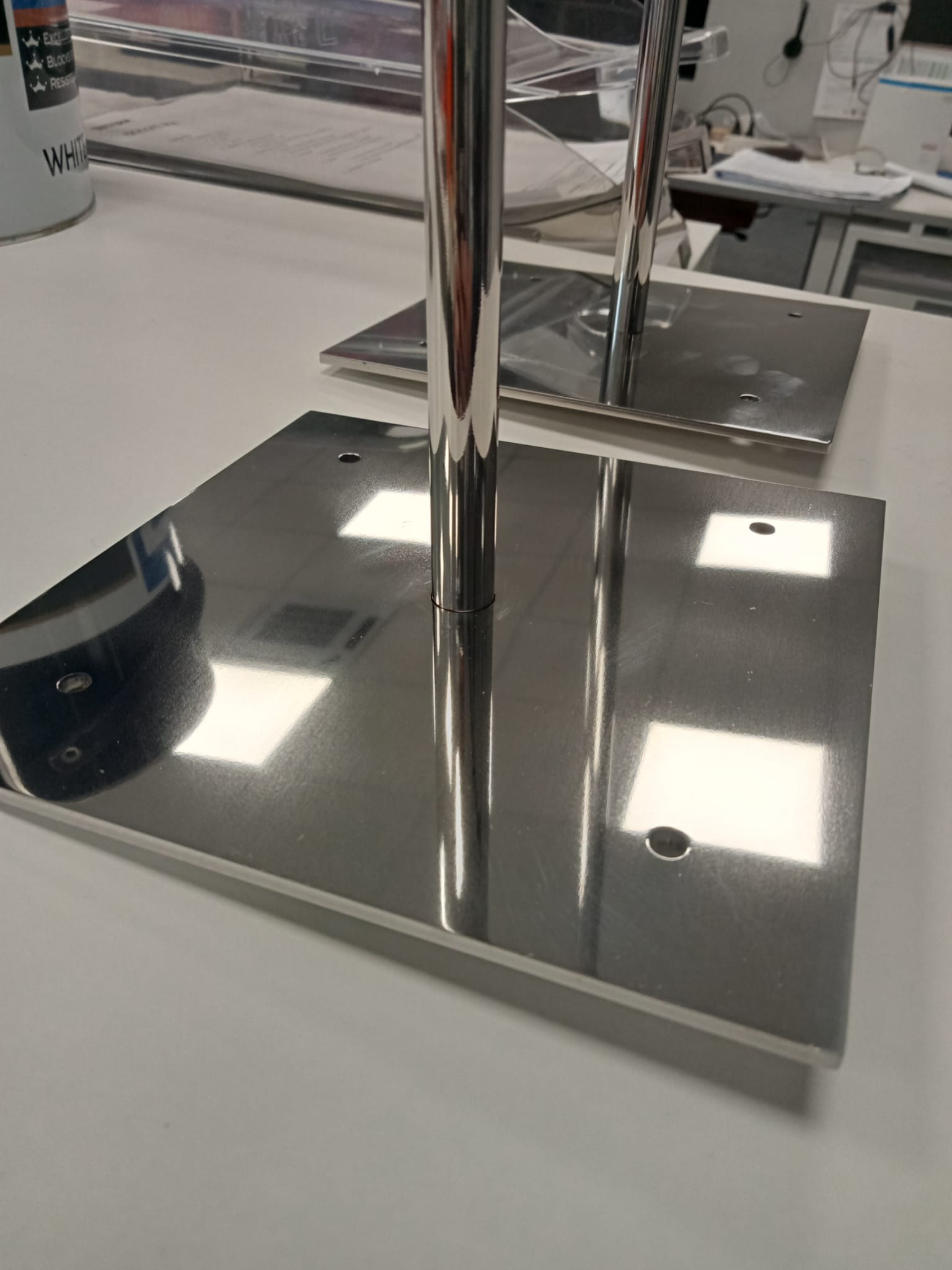 Błyszczące, metalowe słupki z kwadratowymi podstawami, widoczne otwory montażowe, ustawione na jasnym blacie, w tle biurowe otoczenie.