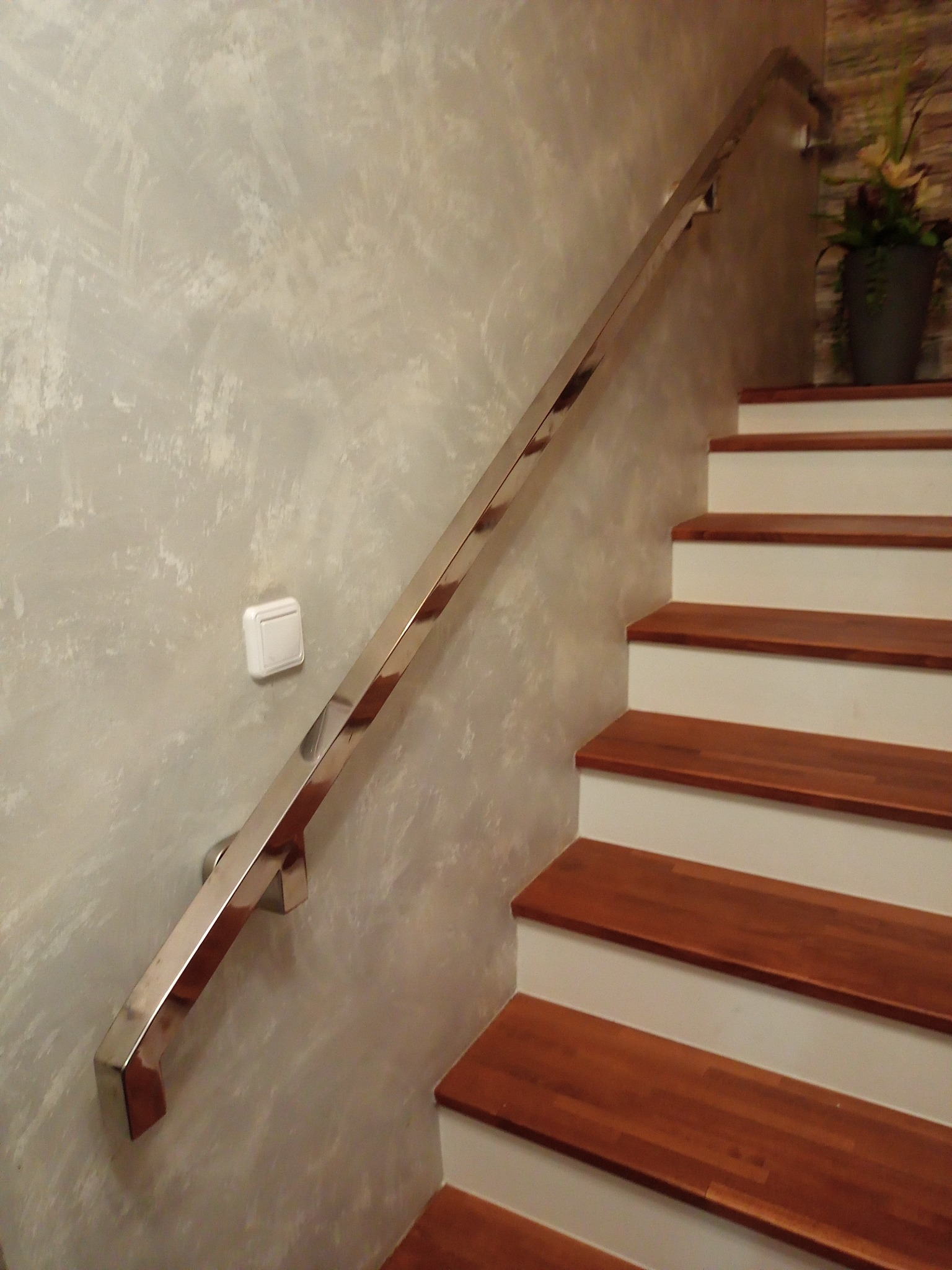 Polerowana metalowa poręcz zamocowana na ścianie przy drewnianych schodach z białymi podstopnicami, widok z dołu, dekoracyjna ściana w tle.