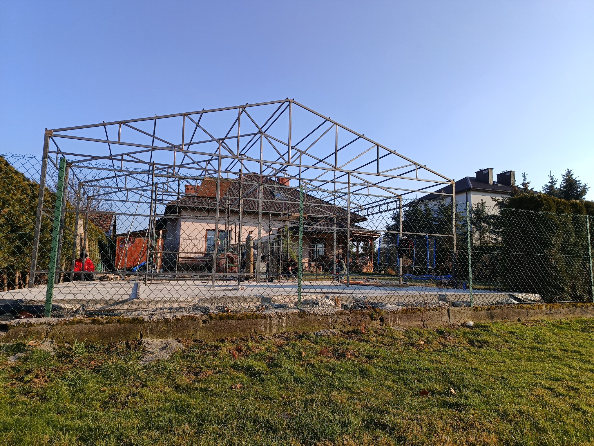 Stalowa konstrukcja z profili, prawdopodobnie zadaszenia tarasu lub wiaty, na tle domu jednorodzinnego i zielonego trawnika, widoczna przez siatkę ogrodzeniową.