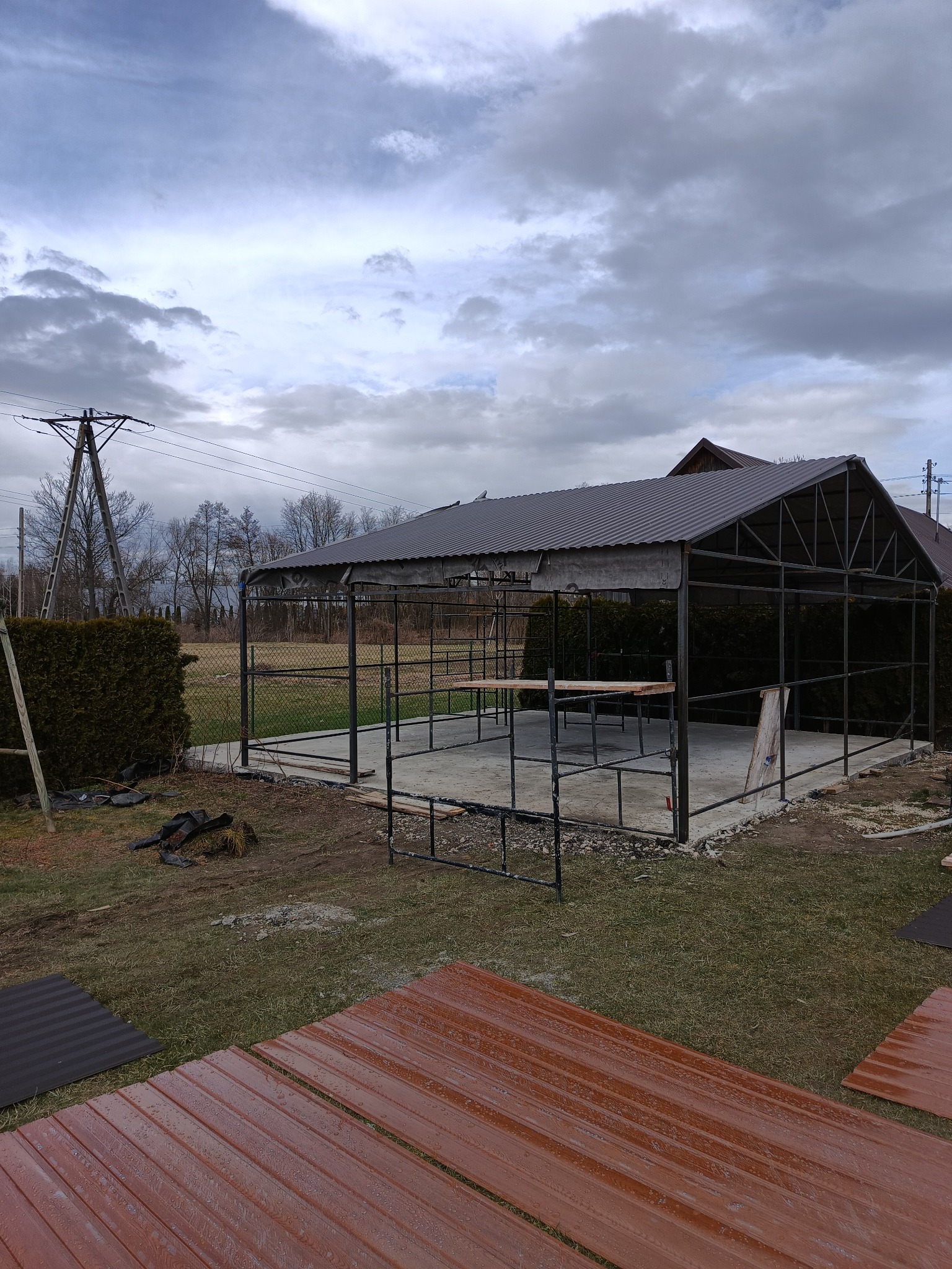 Konstrukcja altany ogrodowej ze stalowych profili w trakcie montażu dachu z blachy trapezowej, widoczne elementy podtrzymujące konstrukcję dachu oraz przygotowane arkusze blachy na pierwszym planie.