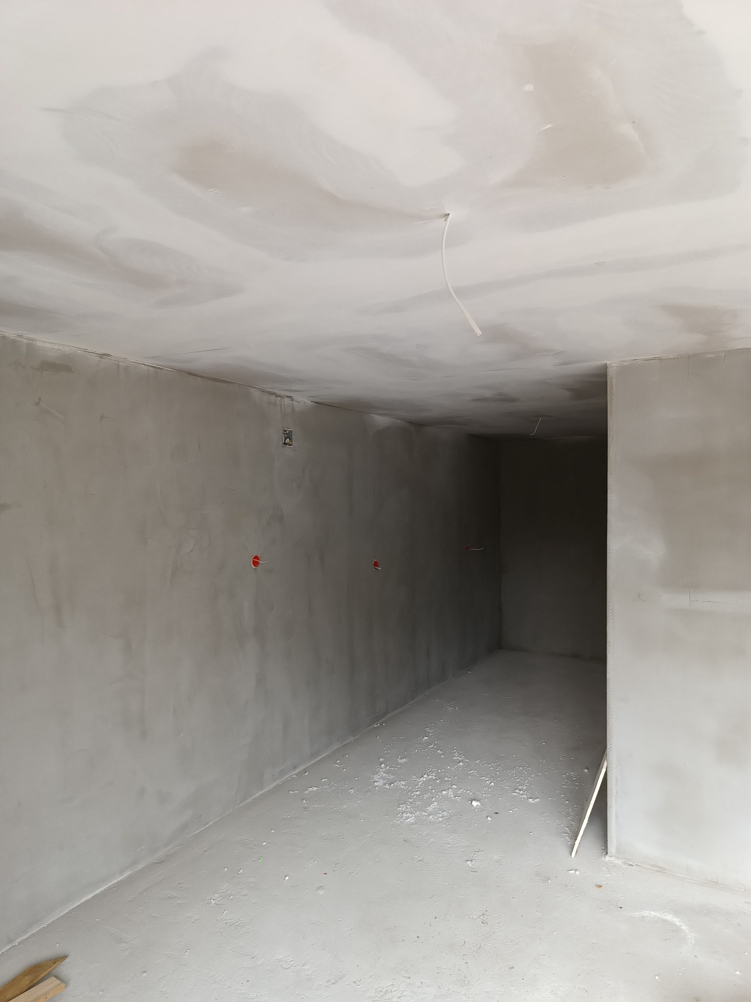 Surowe wnętrze w trakcie wykańczania, widoczne szpachlowane ściany z wyprowadzonymi przewodami elektrycznymi, sufit w trakcie malowania, podłoga z resztkami materiałów budowlanych.