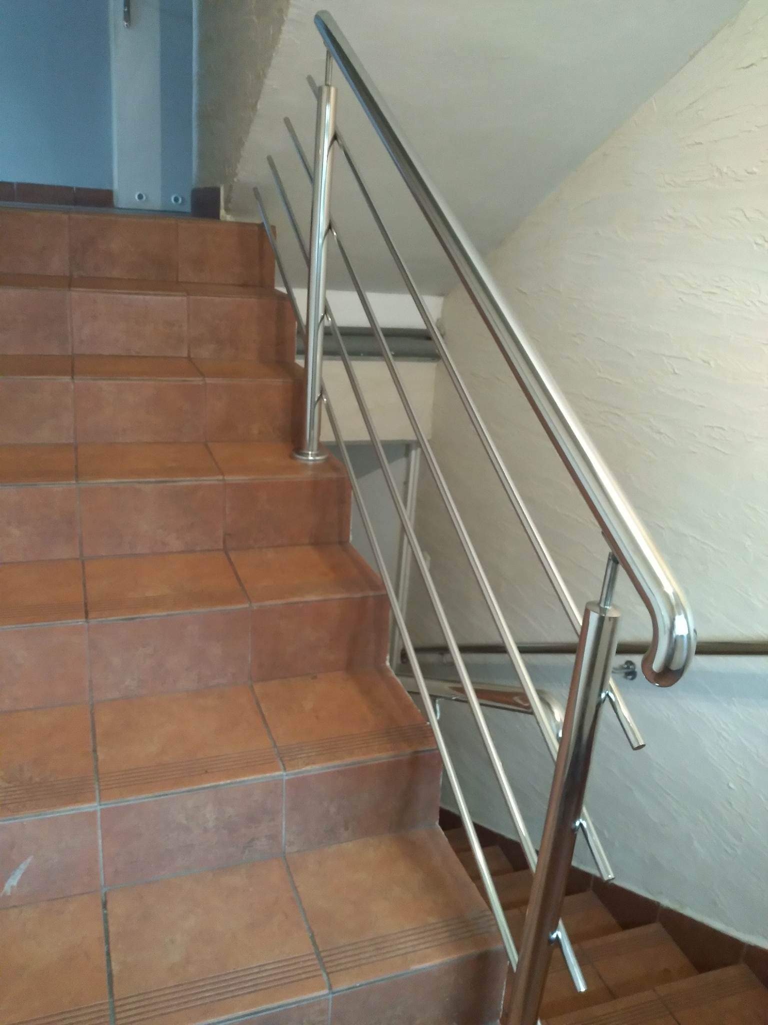 Polerowana, metalowa barierka schodowa z czterema poziomymi prętami, zamontowana na brązowych, ceramicznych stopniach w budynku.