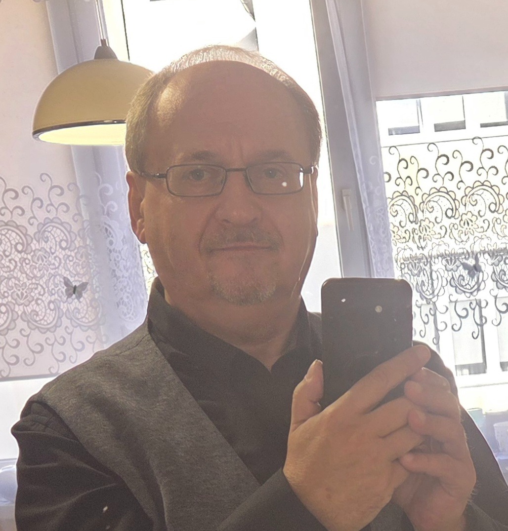 Mężczyzna w okularach robi selfie telefonem na tle okna z firanką. Widoczna lampa sufitowa. Ubrany w koszulę i kamizelkę. Uśmiechnięty, z lekkim zarostem.