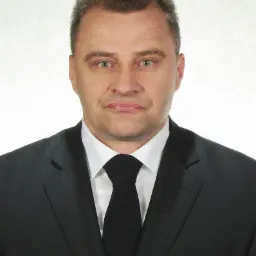 Portret mężczyzny w czarnym garniturze i krawacie na jasnym tle, z widoczną białą poszetką w kieszeni marynarki.