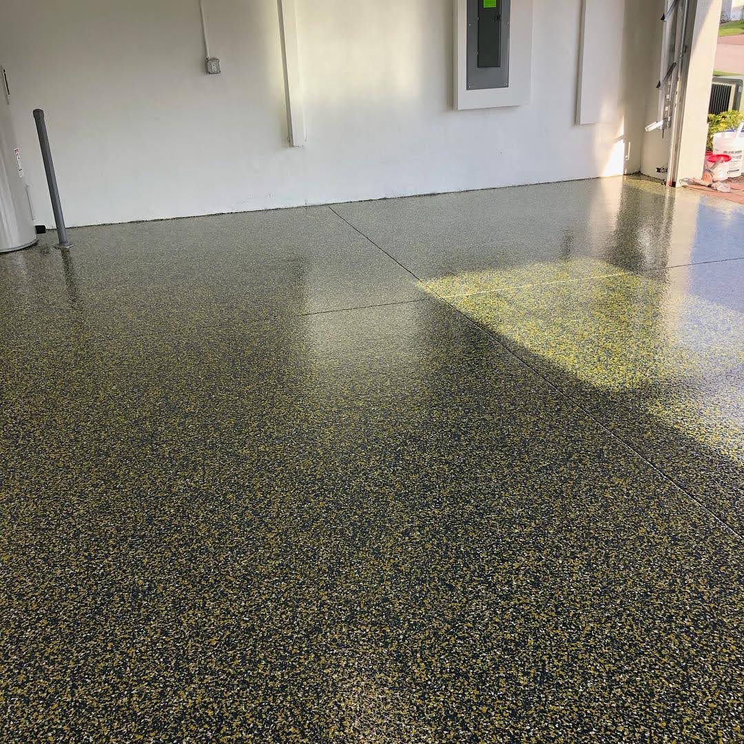 Błyszcząca, bezszwowa podłoga w garażu z drobnymi, czarno-złotymi plamkami, odbijająca światło słoneczne wpadające przez otwarte drzwi.