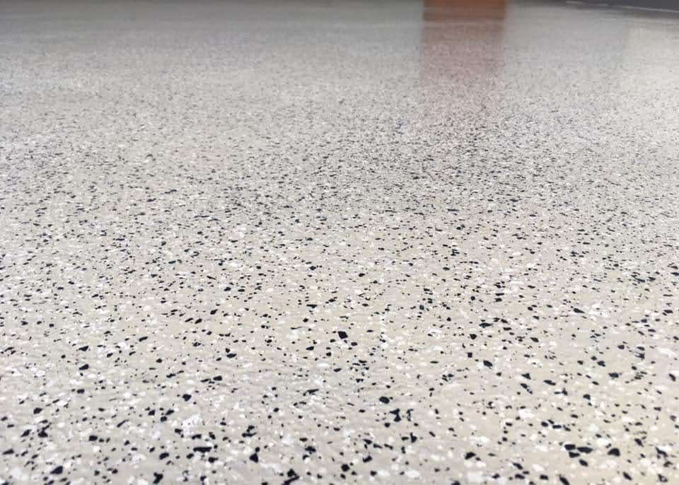 Jasnoszara, gładka powierzchnia z czarnymi i białymi drobinkami, przypominająca lastryko, odbijająca górne światło.