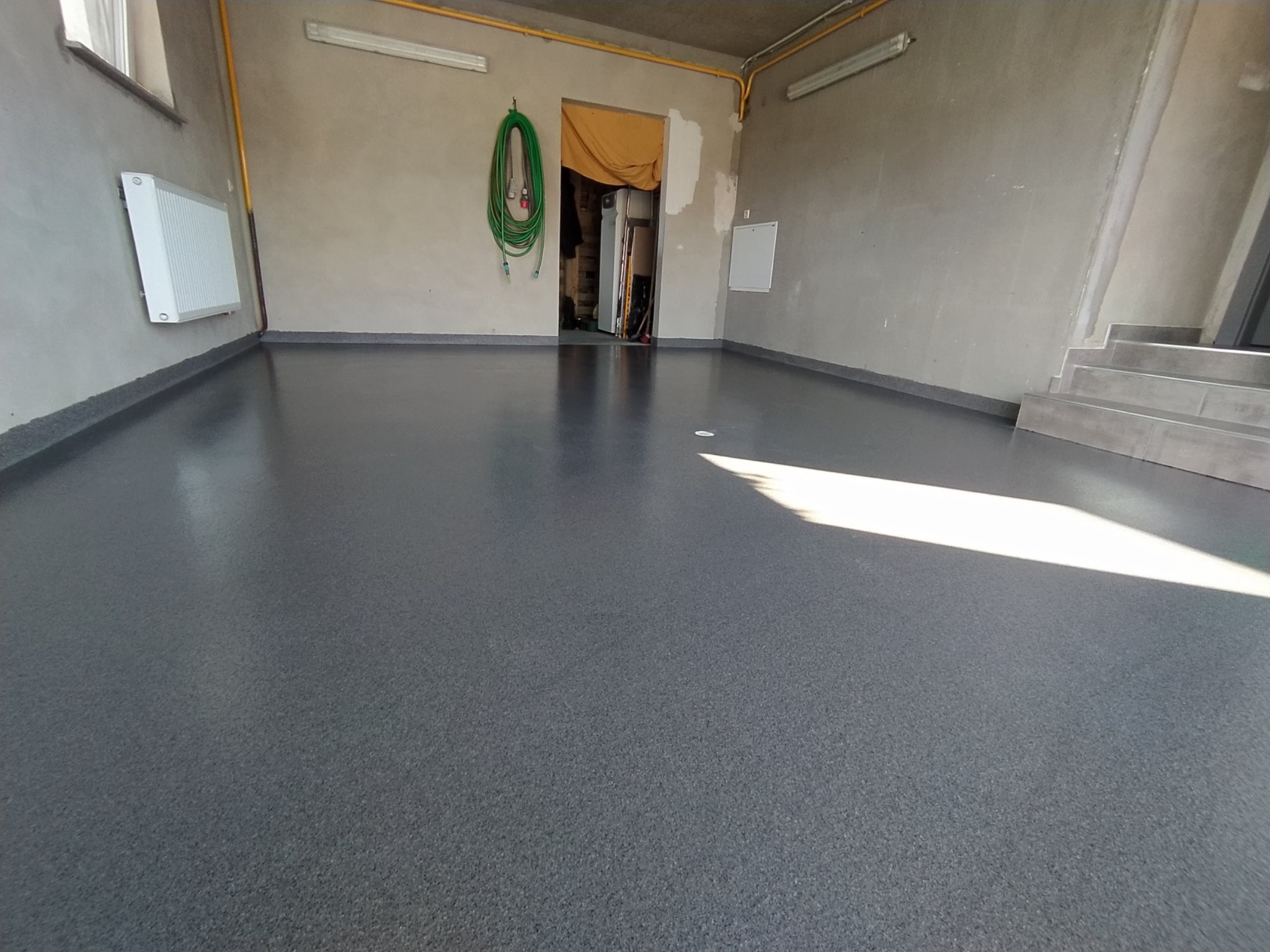 Szara, gładka posadzka w pomieszczeniu z betonowymi ścianami, widoczny grzejnik, zwinięty wąż ogrodowy i schody w tle.