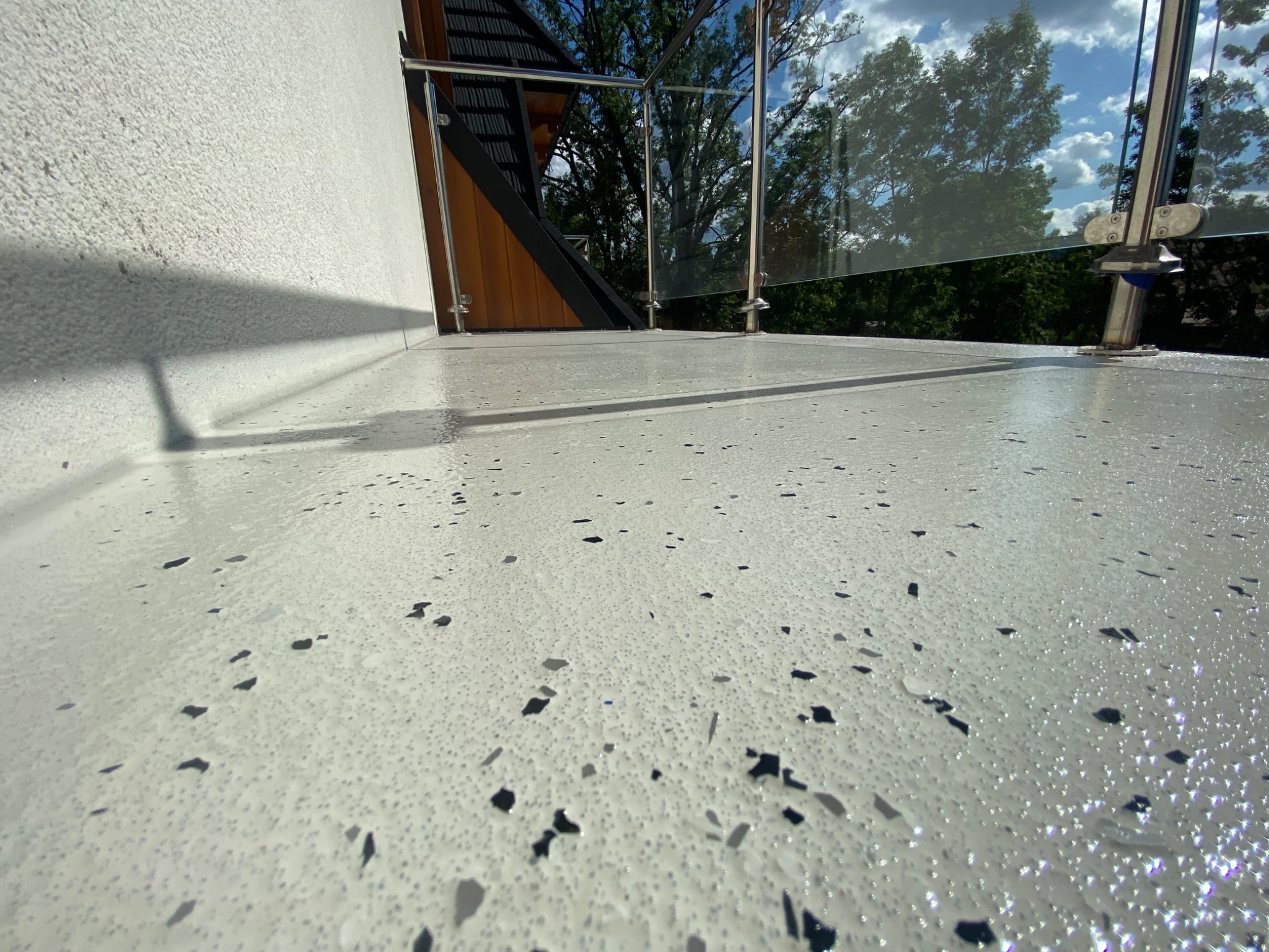 Jasnoszara powierzchnia balkonu z drobnymi czarnymi elementami dekoracyjnymi, szklana balustrada z metalowymi mocowaniami, widok na drzewa i niebo w tle.