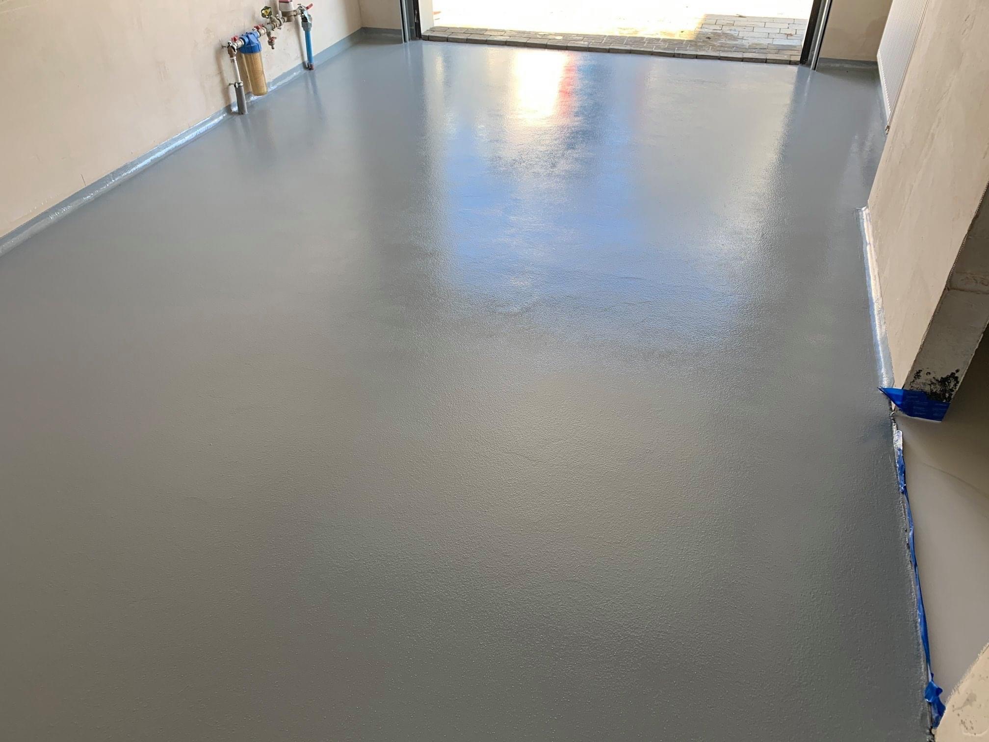 Świeżo wylana, lśniąca szara podłoga w pomieszczeniu z widocznymi rurami i filtrami przy ścianie, z delikatnym odbiciem światła w centralnej części.