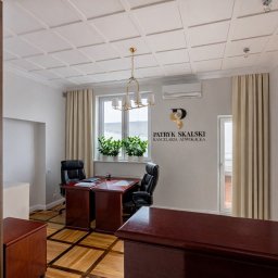 Kancelaria Adwokacka Adwokat Patryk Skalski - Eleganckie wnętrze kancelarii adwokackiej z drewnianymi meblami, parkietem i logo na ścianie. Widok na biurko, krzesła i okno z roślinami. Oświetlenie sufitowe.
