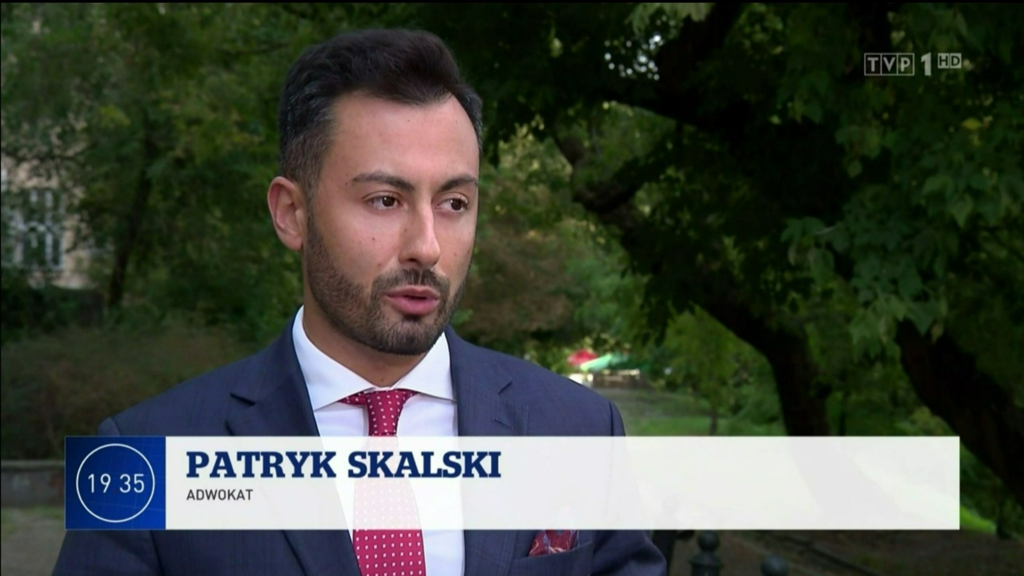 Mężczyzna w garniturze i krawacie w kropki, udzielający wywiadu w plenerze, z widocznym logo TVP1 HD w rogu ekranu.