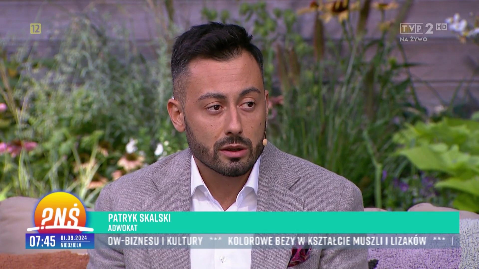 Portret mężczyzny w jasnej marynarce, prawdopodobnie adwokata Patryka Skalskiego, podczas wywiadu w programie telewizyjnym na tle roślin.