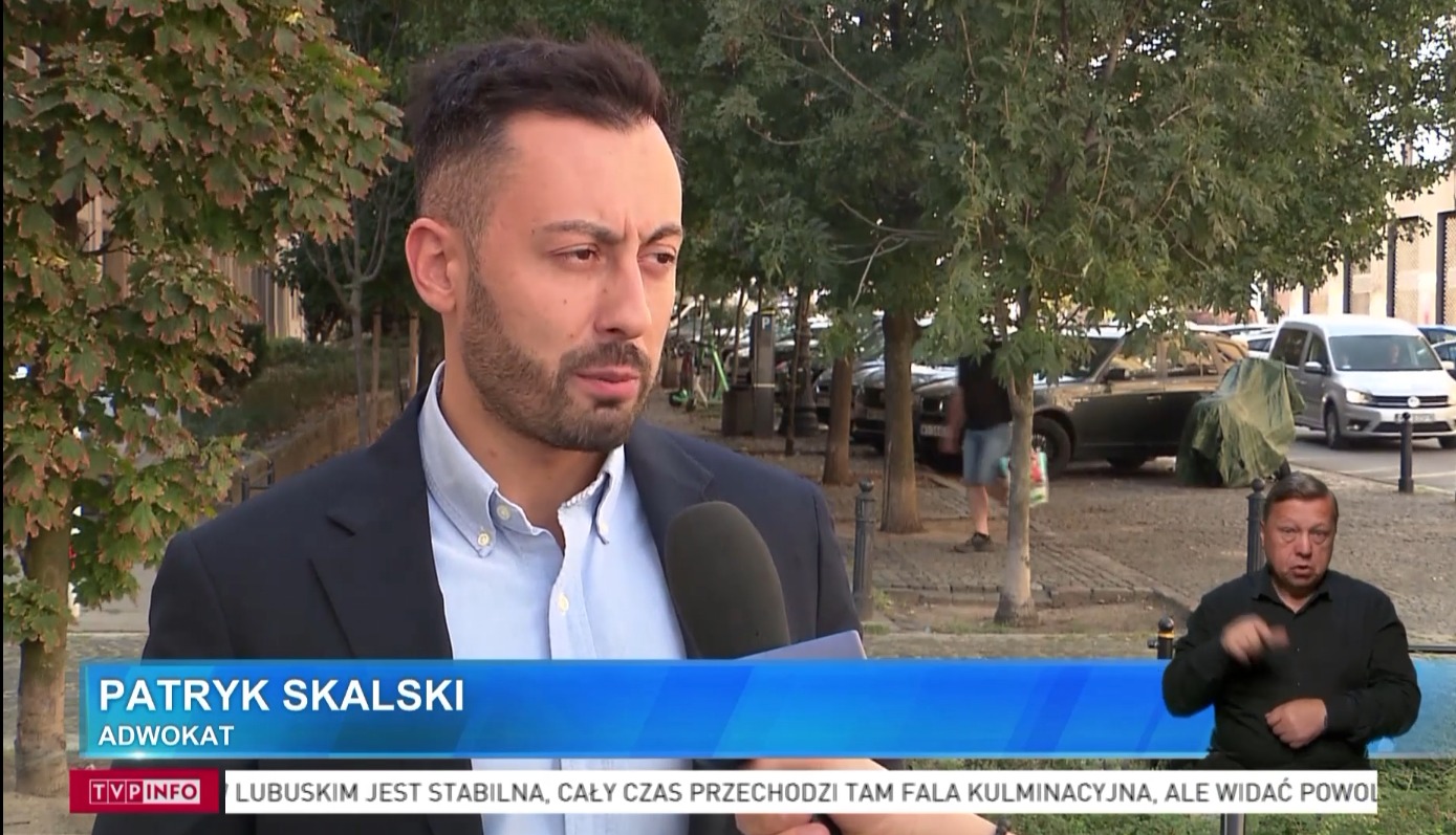 Mężczyzna z brodą w garniturze udziela wywiadu do mikrofonu, w tle drzewa i samochody na ulicy.